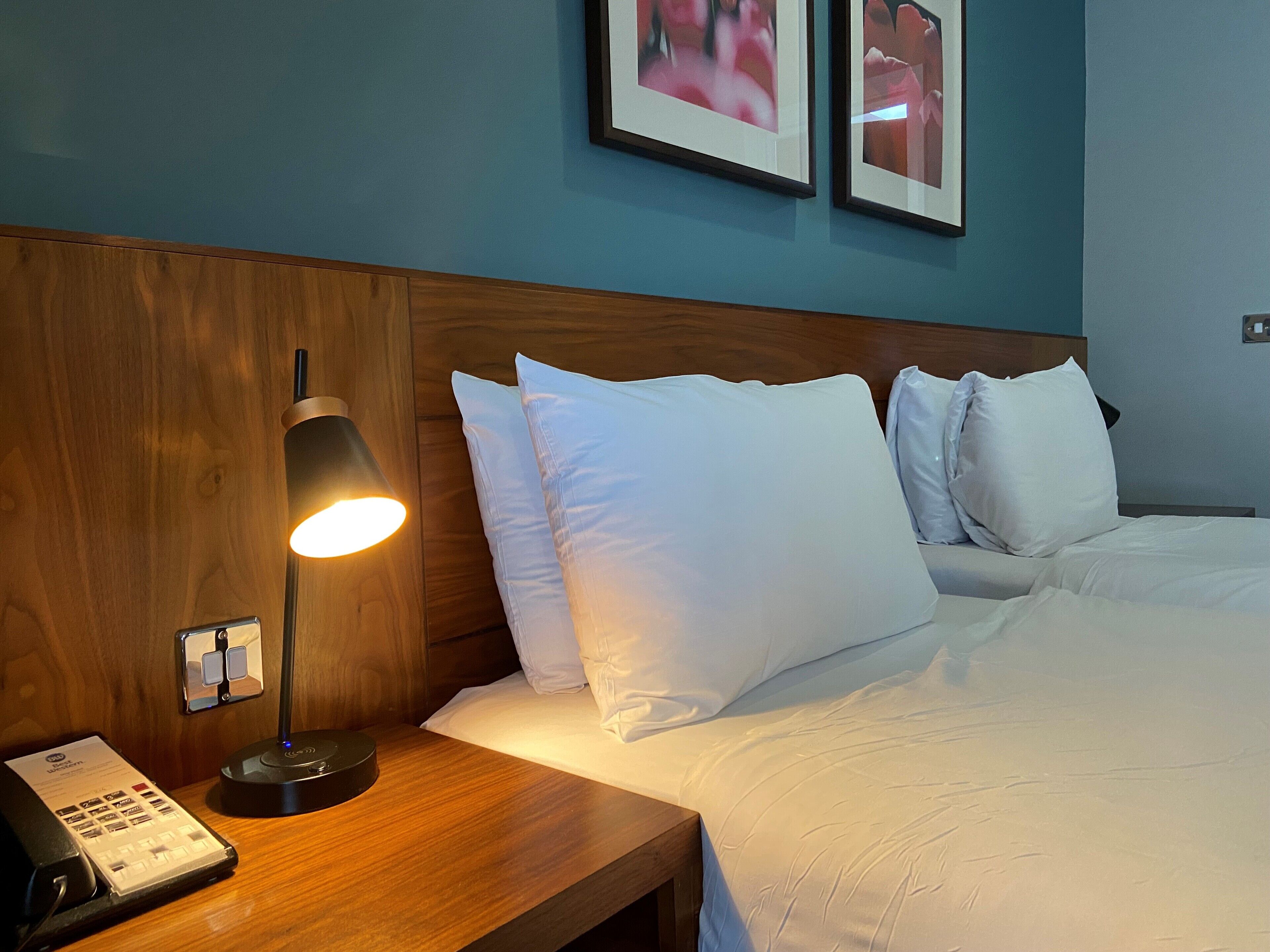 Ett hotellrum med en sänggavel i trä, vita sängkläder, en sänglampa, en telefon och två inramade tavlor på väggen.