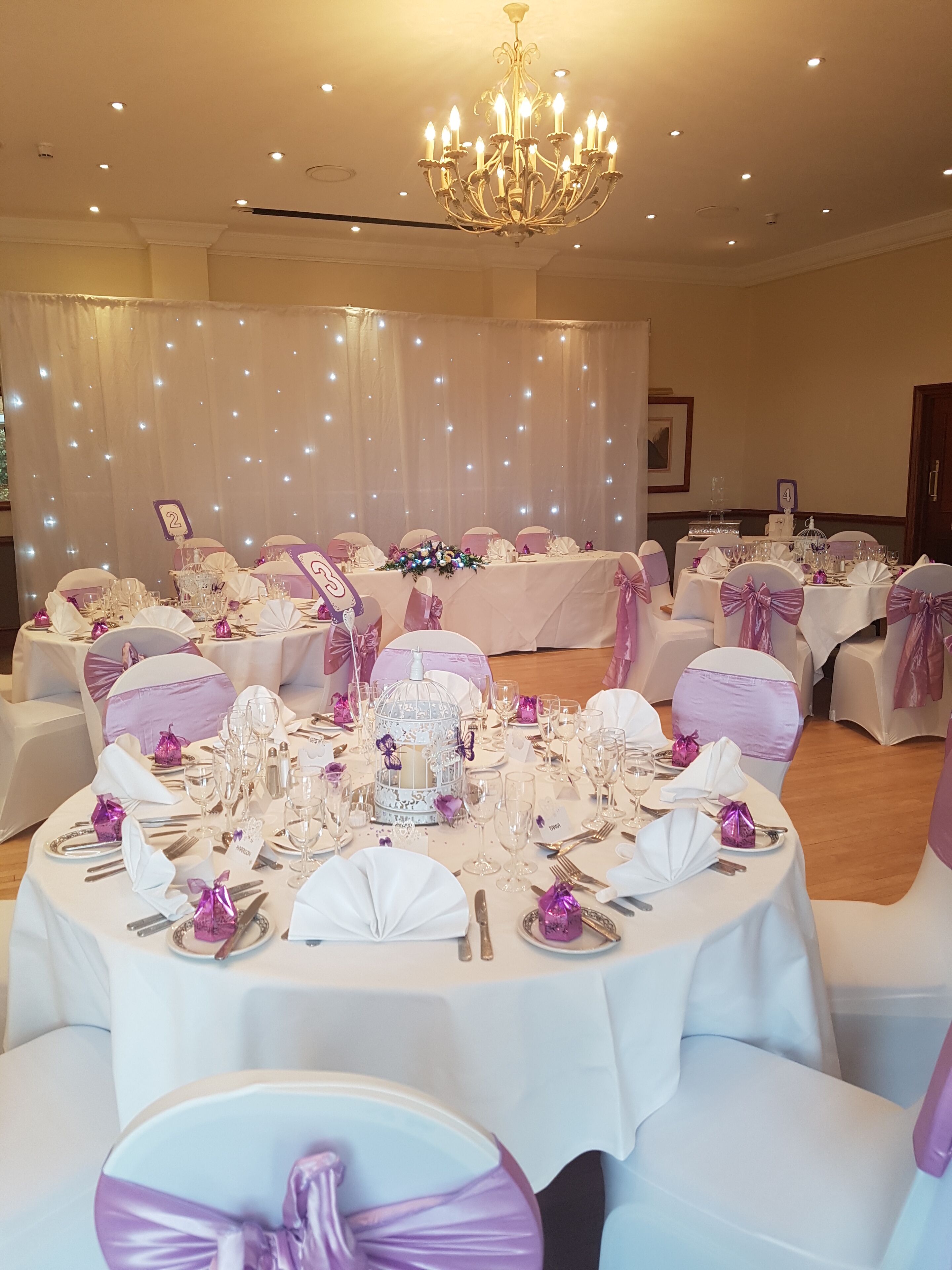 A banquet hall with round tables set for a formal event, featuring white tablecloths, purple sashes, and a chandelier.