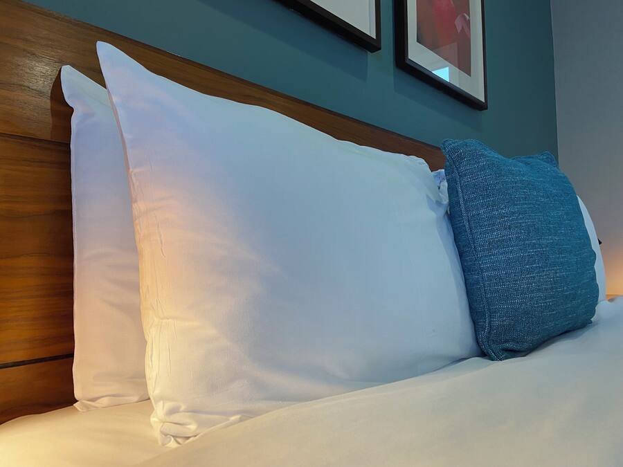Una cama con almohadas blancas y azules, cabecera de madera y pared verde azulada con cuadros.