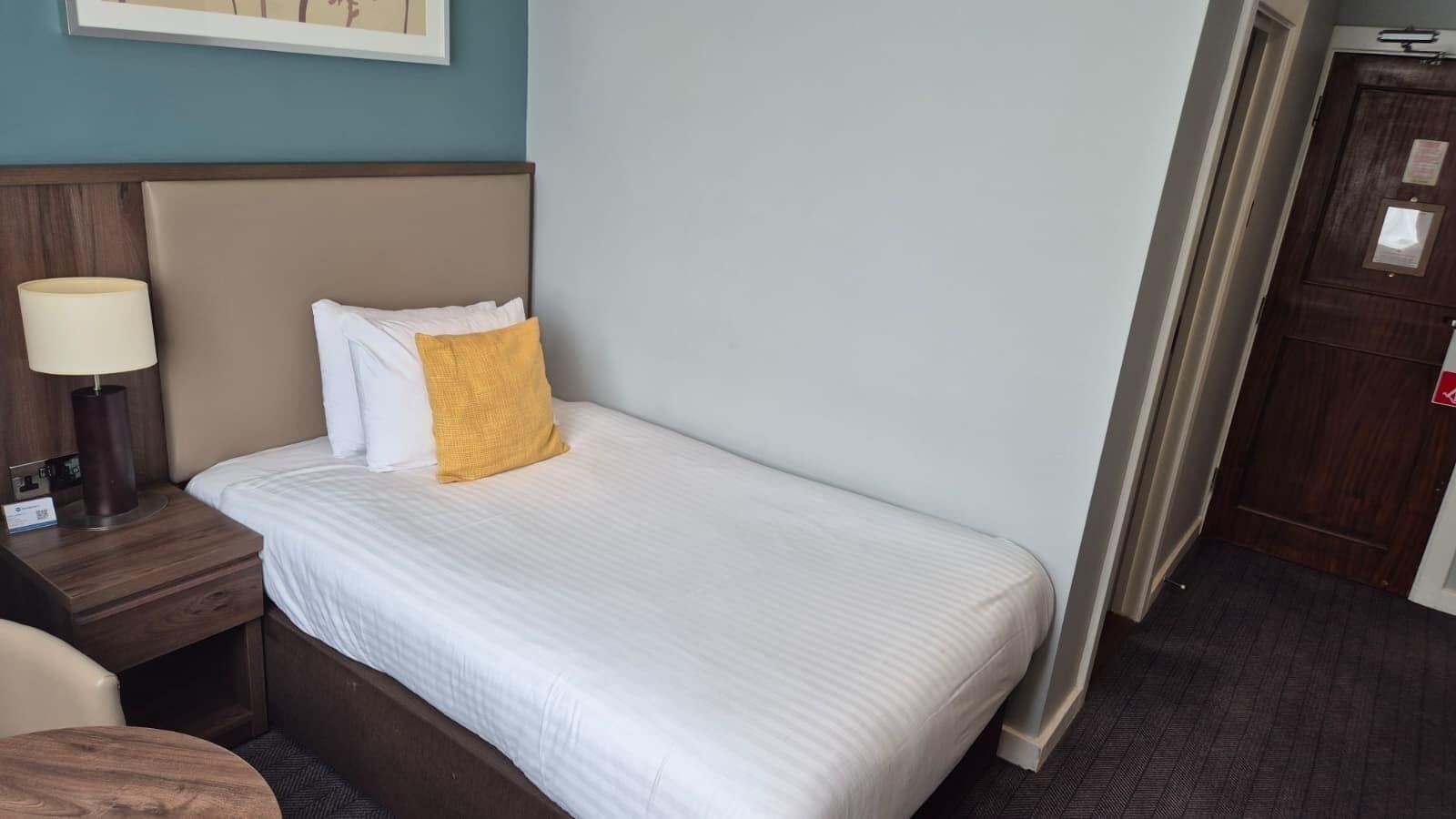 A hotel room with a bed, a nightstand, a lamp, and a door.