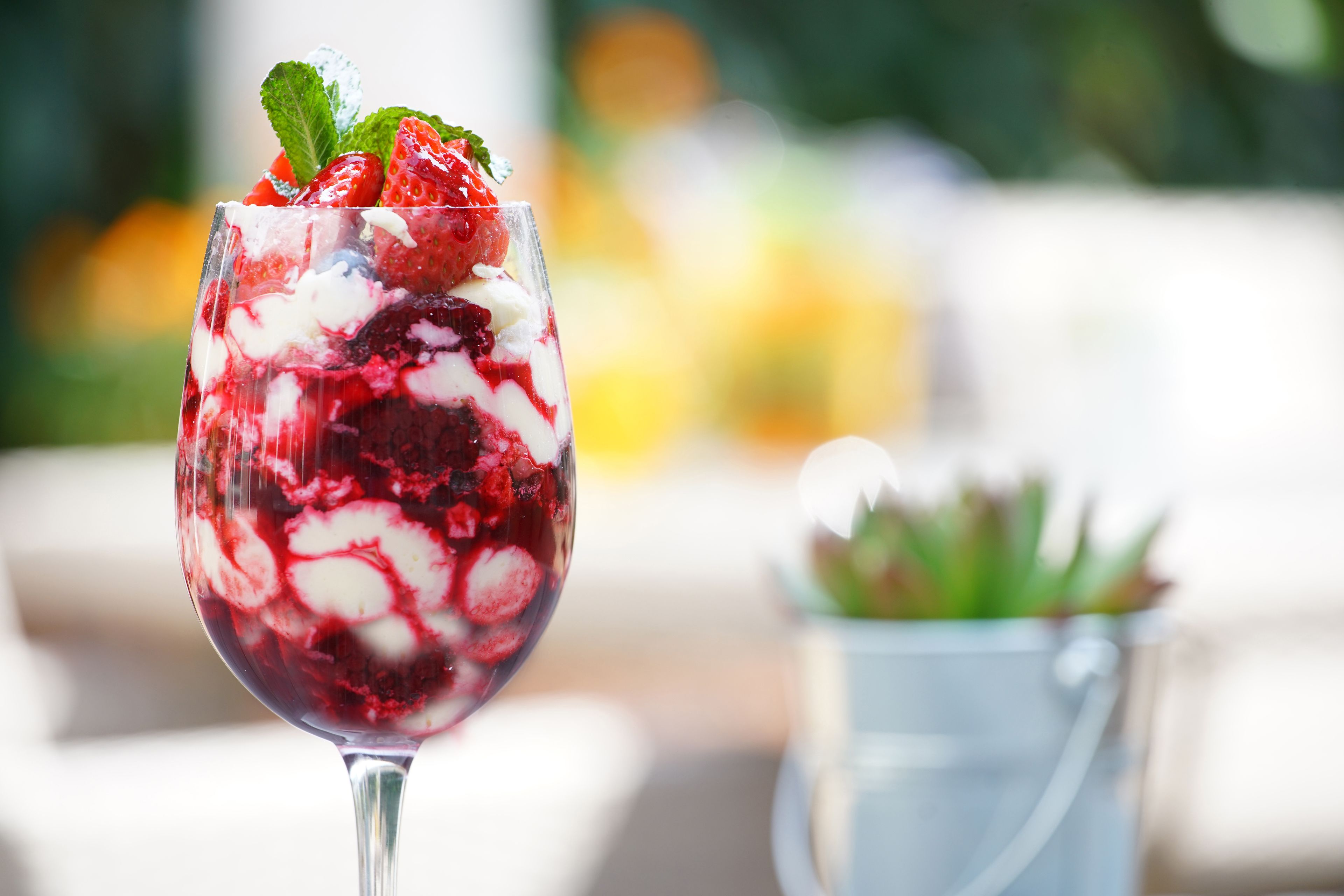 A dessert in a glass with strawberries and mint, with a blurred background of greenery and a white object.