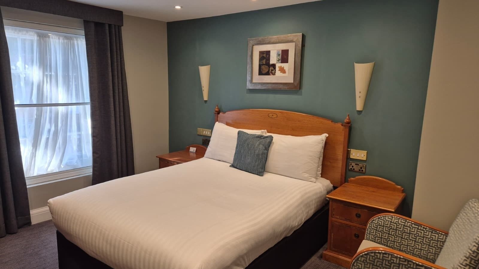 Habitació d'hotel amb un llit gran, una tauleta de nit, una cadira, una finestra amb cortines i un quadre penjat a la paret.