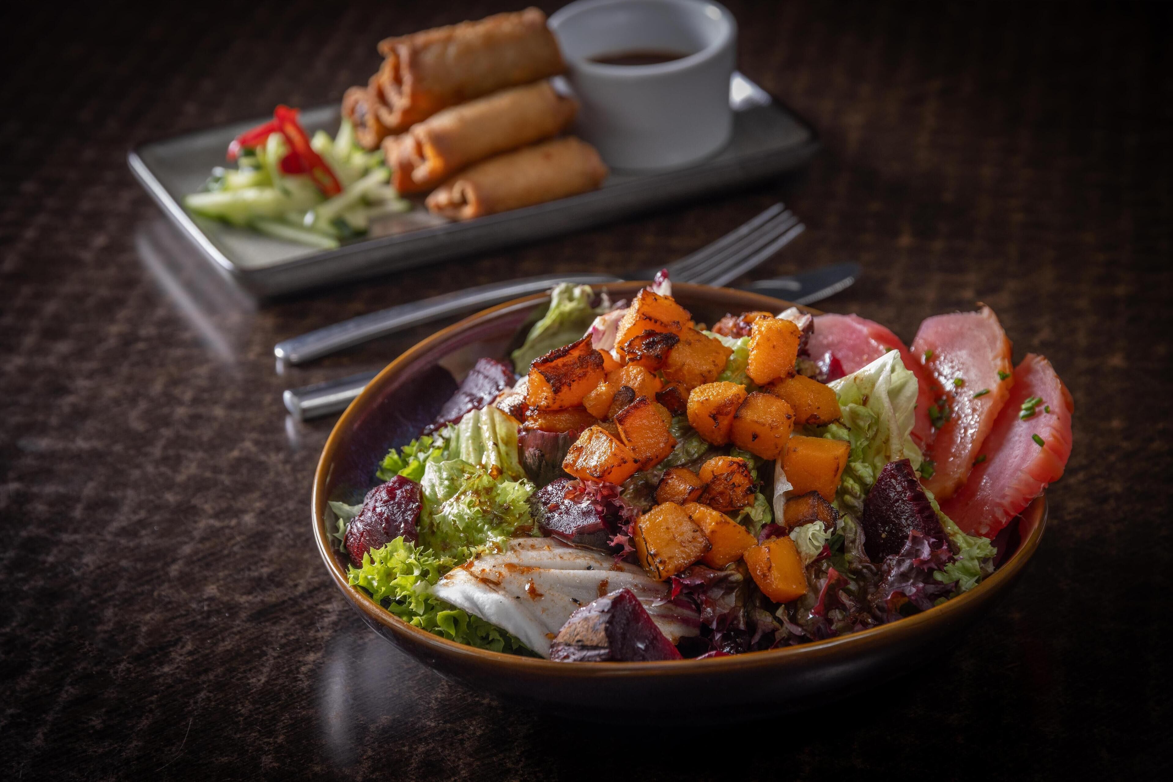 A bowl of salad with grilled vegetables and meat, a plate of spring rolls, and a small cup of sauce on a dark table.