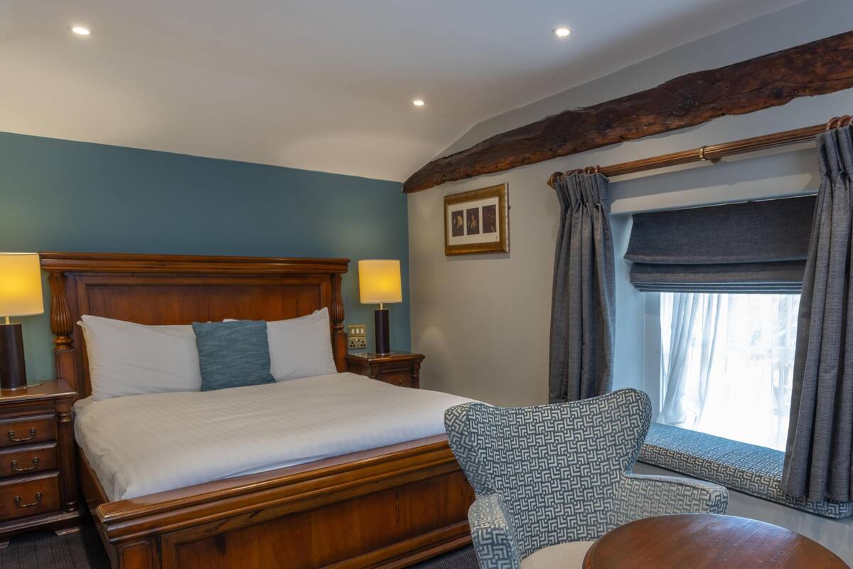 A neatly made bed with a wooden headboard, a bedside table with a lamp, a patterned armchair, and a window with curtains.