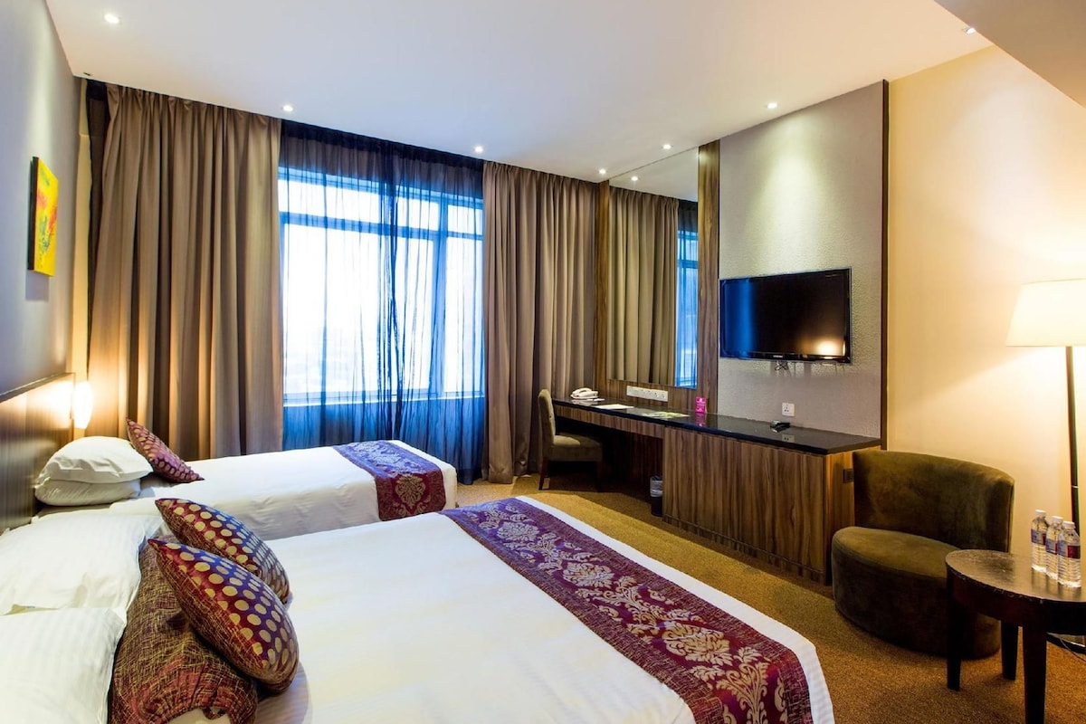 A hotel room with two beds, a desk, a chair, a TV, and a lamp.