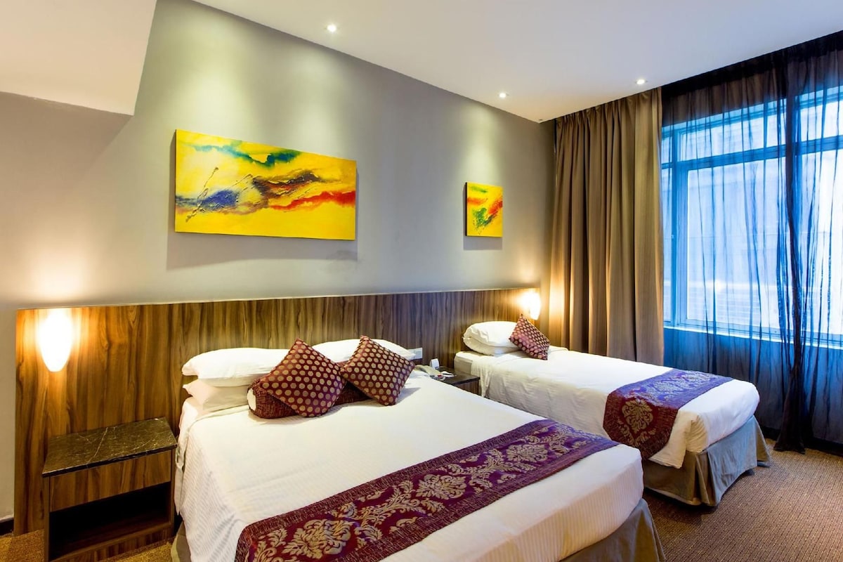 Une chambre d’hôtel avec deux lits, une tête de lit en bois, une grande fenêtre avec des rideaux et deux tableaux abstraits accrochés au mur.