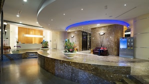 Zona de recepción de hotel con mostrador de mármol, escritorio de recepción, sofá y quiosco informativo digital.
