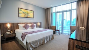 Habitación de hotel con una cama grande, un escritorio, una silla y vistas de la ciudad a través de amplios ventanales.