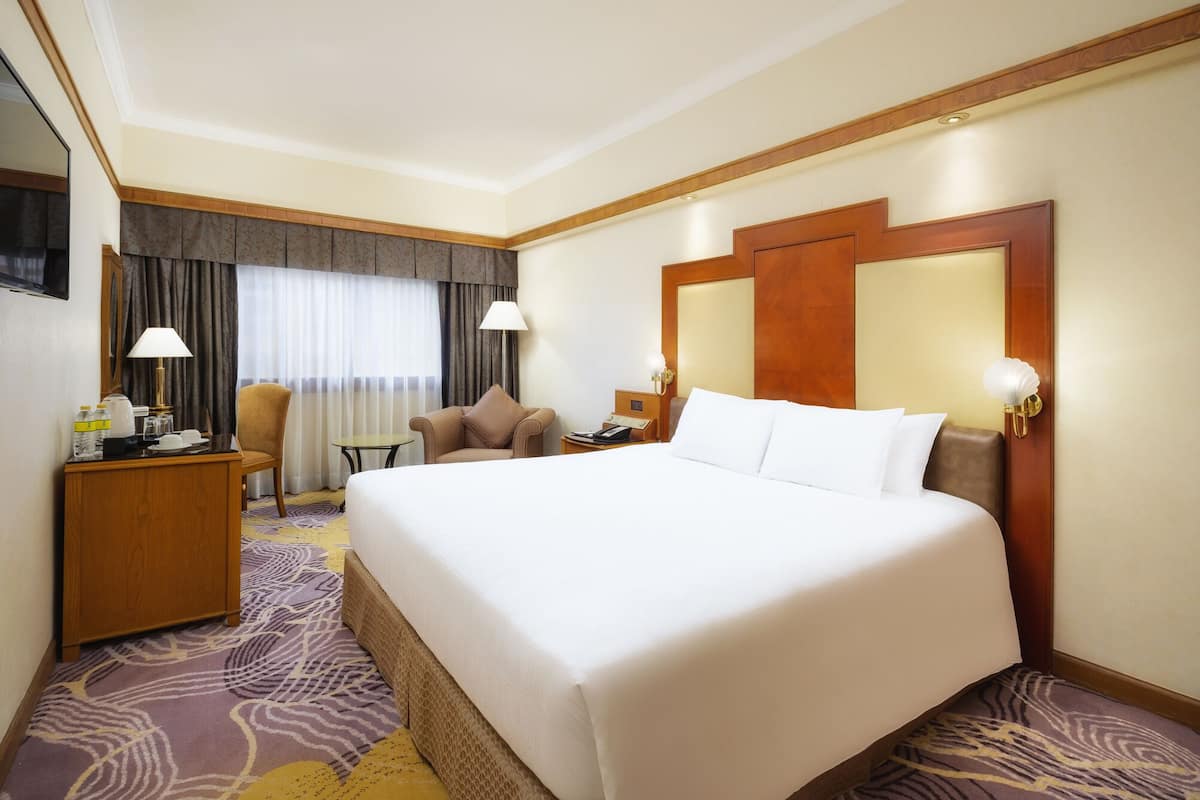 A hotel room with a large bed, a desk with a lamp, a chair, a small table, and a TV on the wall.