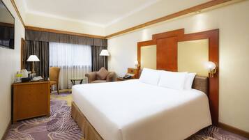 A hotel room with a large bed, a desk with a lamp, a chair, a small table, and a TV on the wall.