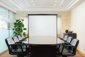 A conference room with a large table, black leather chairs, a projector screen, a potted plant, and a small table with a telephone and flowers. - Hotel Sintra (Macau)