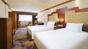 A hotel room with two beds, a wooden wardrobe, a desk, a chair, and a small table with a lamp. - Hotel Sintra (Macau)