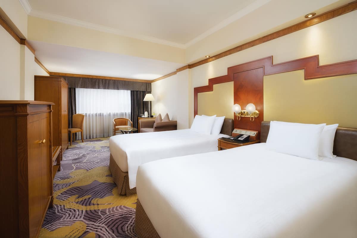 A hotel room with two beds, a wooden wardrobe, a desk, a chair, and a small table with a lamp.