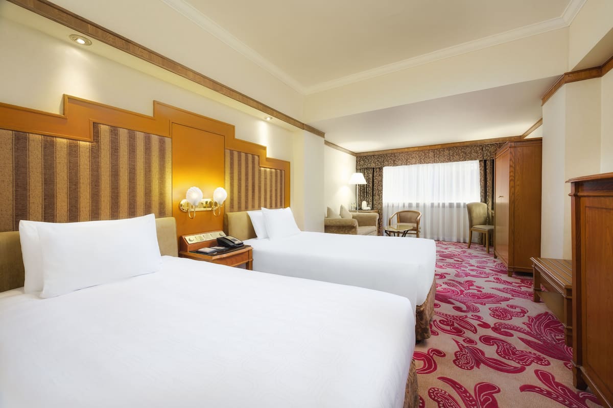 A hotel room with two beds, a desk, a chair, a lamp, and a window with curtains.