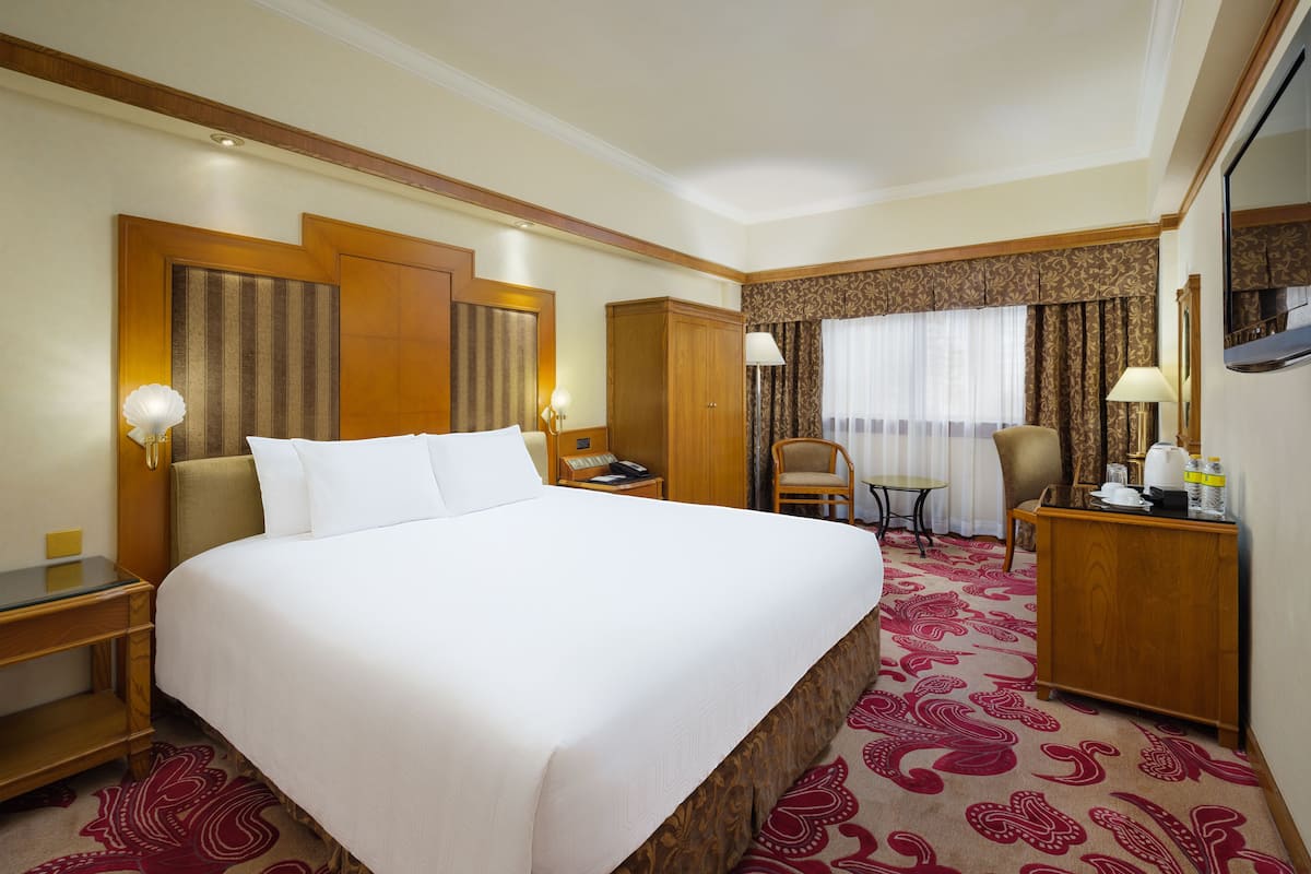 A hotel room with a large bed, a desk, a chair, a small table, and a wardrobe.