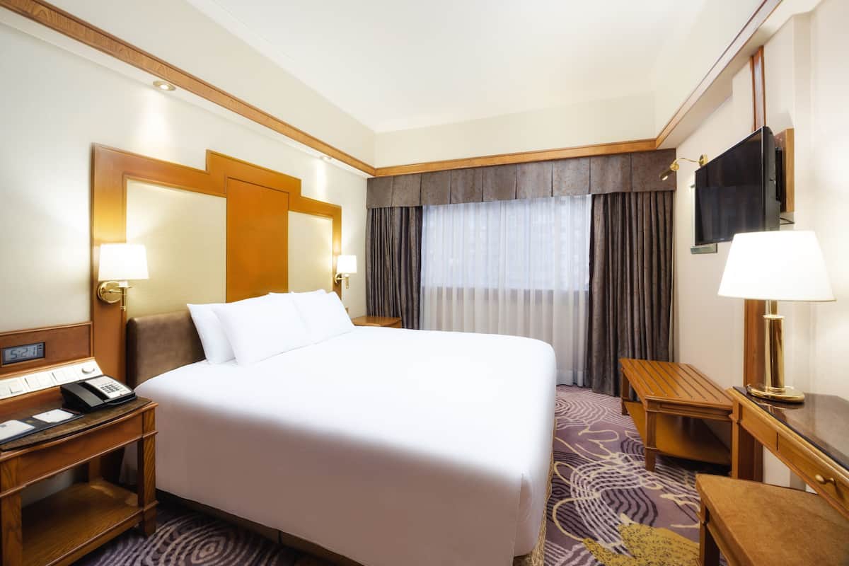 A hotel room with a large bed, bedside tables with lamps, a flat-screen TV mounted on the wall, and a window with curtains.