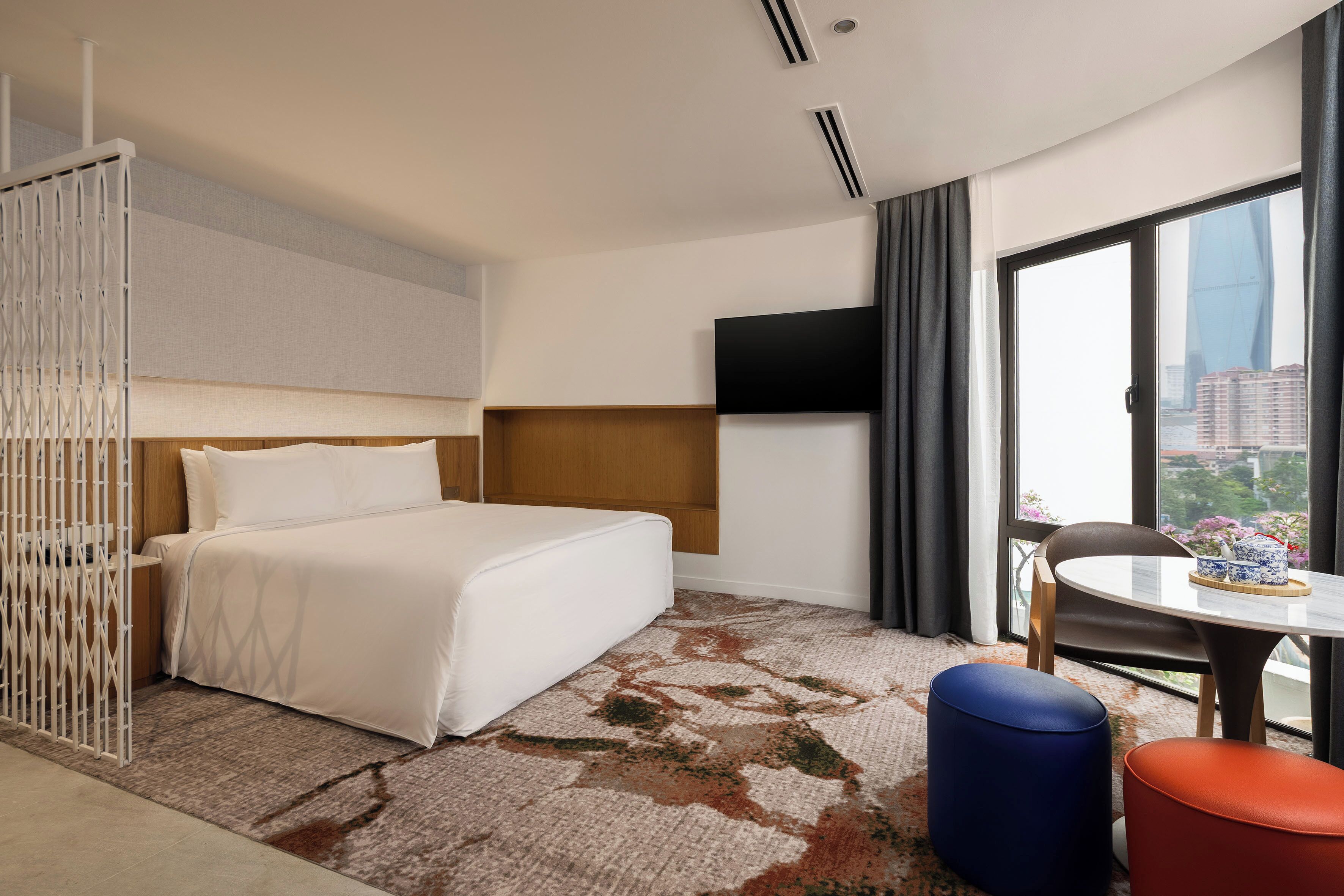 Habitación de hotel con cama, televisión, ventana con vista a la ciudad y una pequeña mesa con un florero.