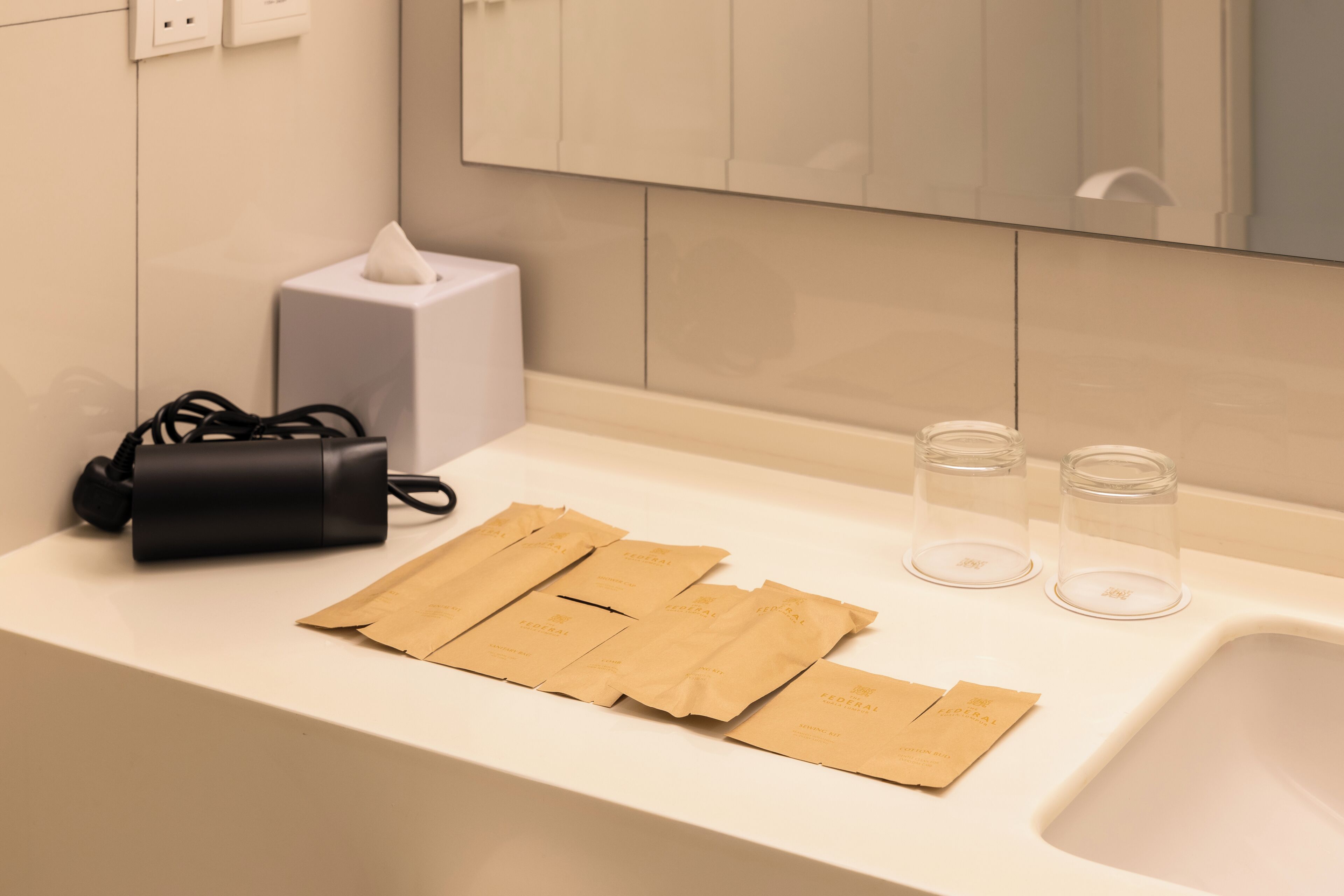 Un baño con encimera blanca, un objeto cilíndrico negro, dos vasos transparentes y varias bolsas de papel marrón.