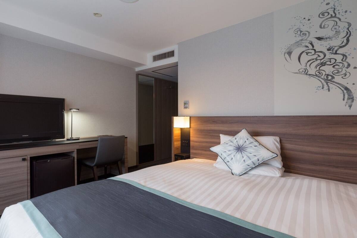 A hotel room with a bed, a desk with a television, a chair, and a nightstand with a lamp.