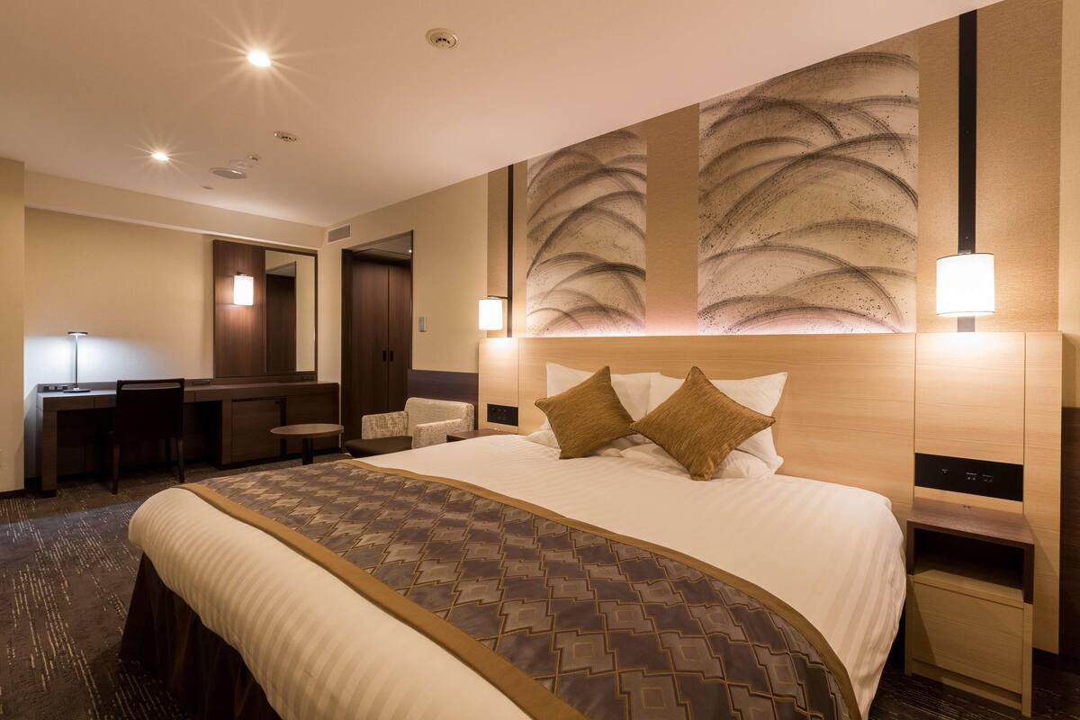 A hotel room with a large bed, a desk, a chair, a small table, and a mirror.