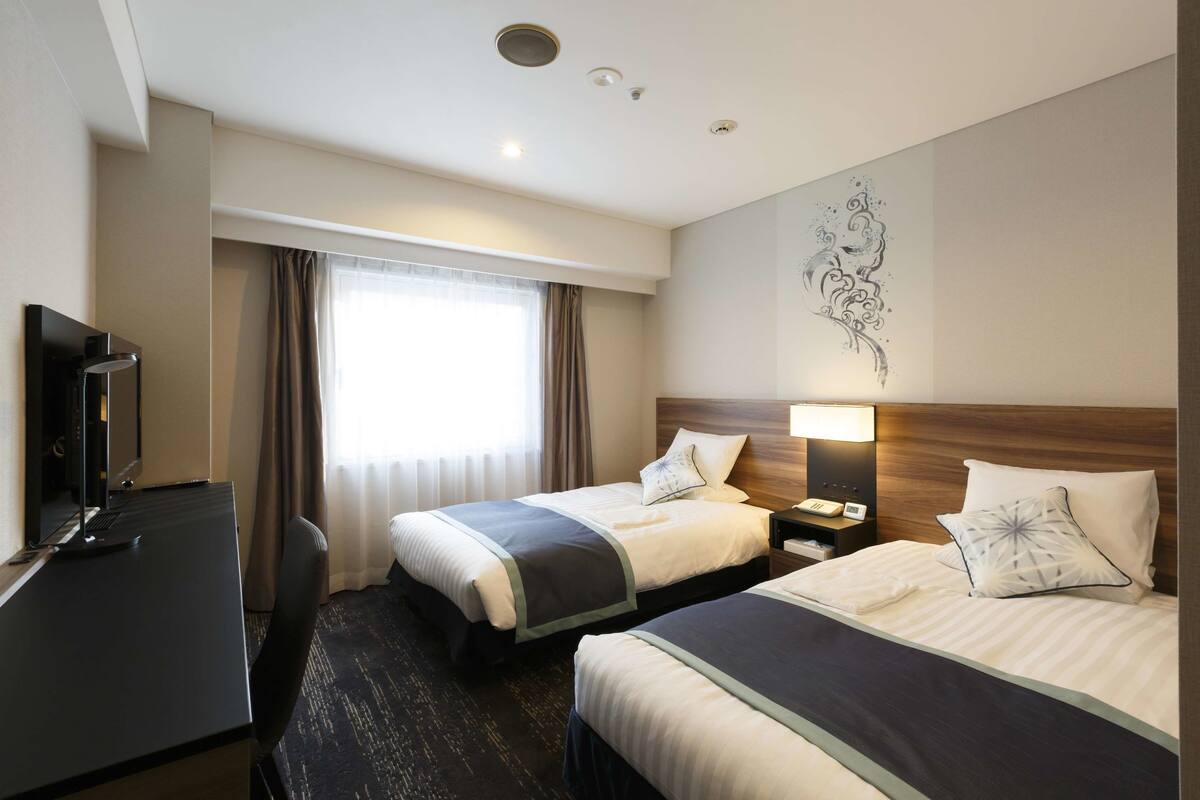 A hotel room with two beds, a desk, a TV, and a window with curtains.