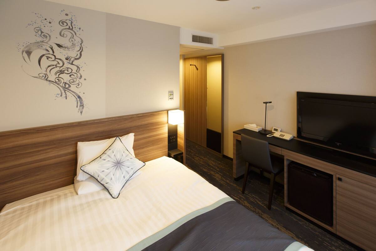 A hotel room with a bed, a desk with a television, a chair, and a door leading to another room.