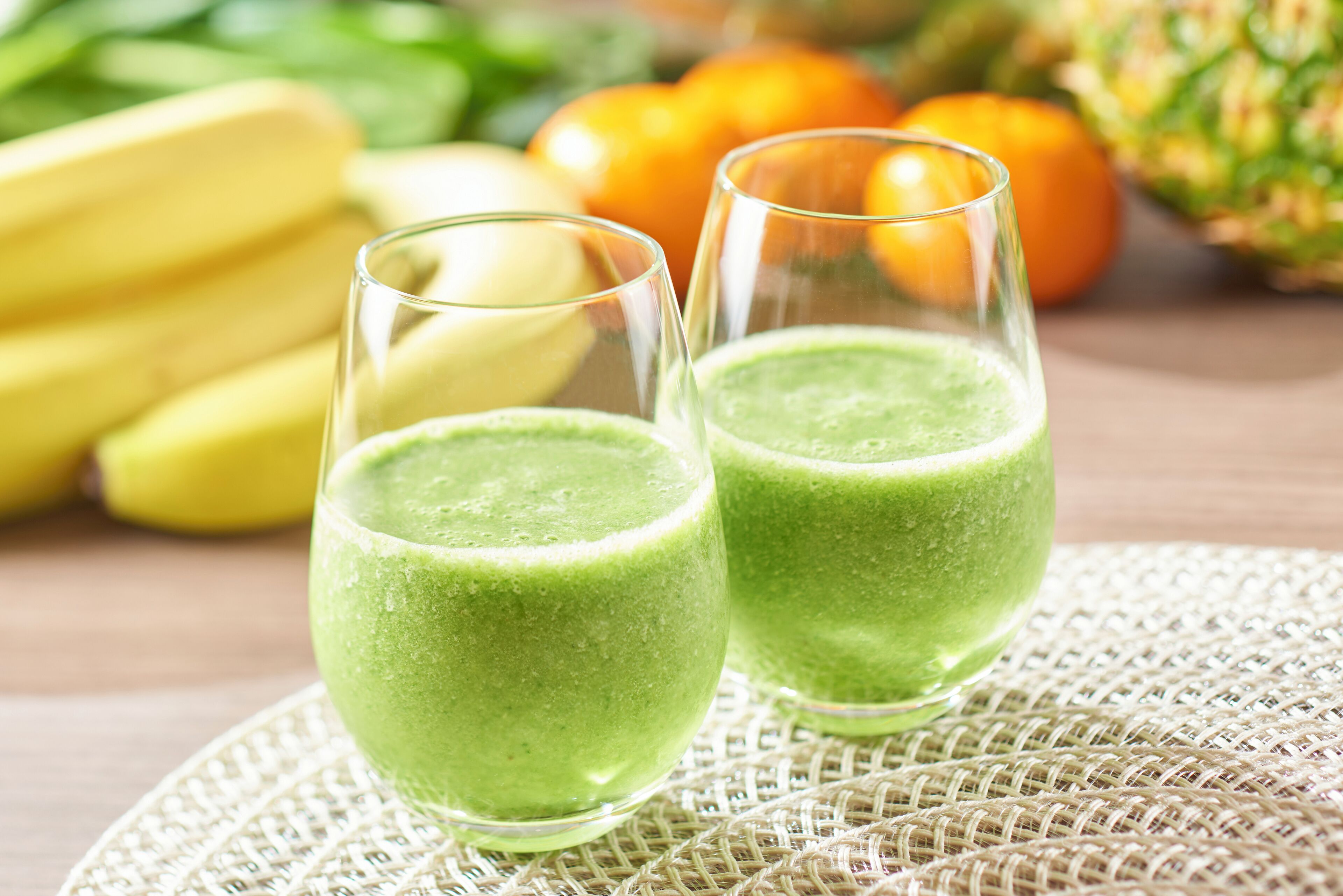 Dos vasos de batido verde sobre un posavasos tejido, con plátanos y naranjas al fondo.