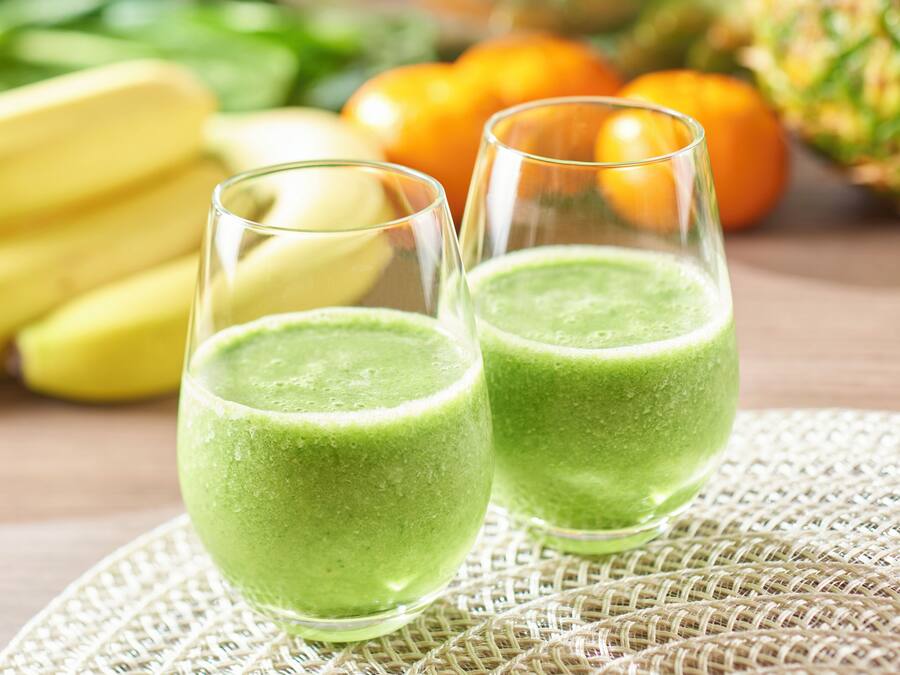 Dos vasos de batido verde sobre un posavasos tejido, con plátanos y naranjas al fondo.