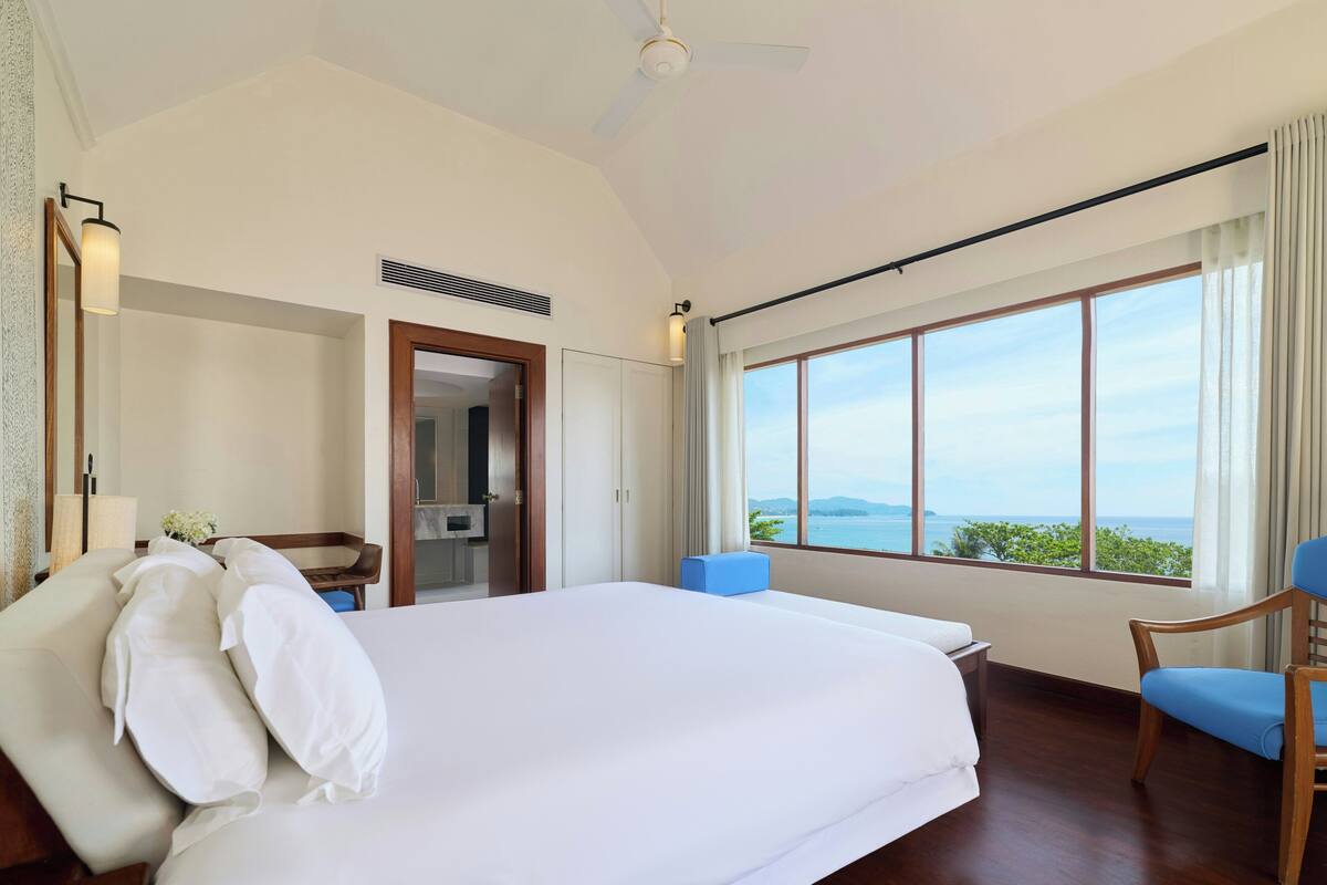 A bedroom with a large bed, a chair, a desk, and a view of the ocean.