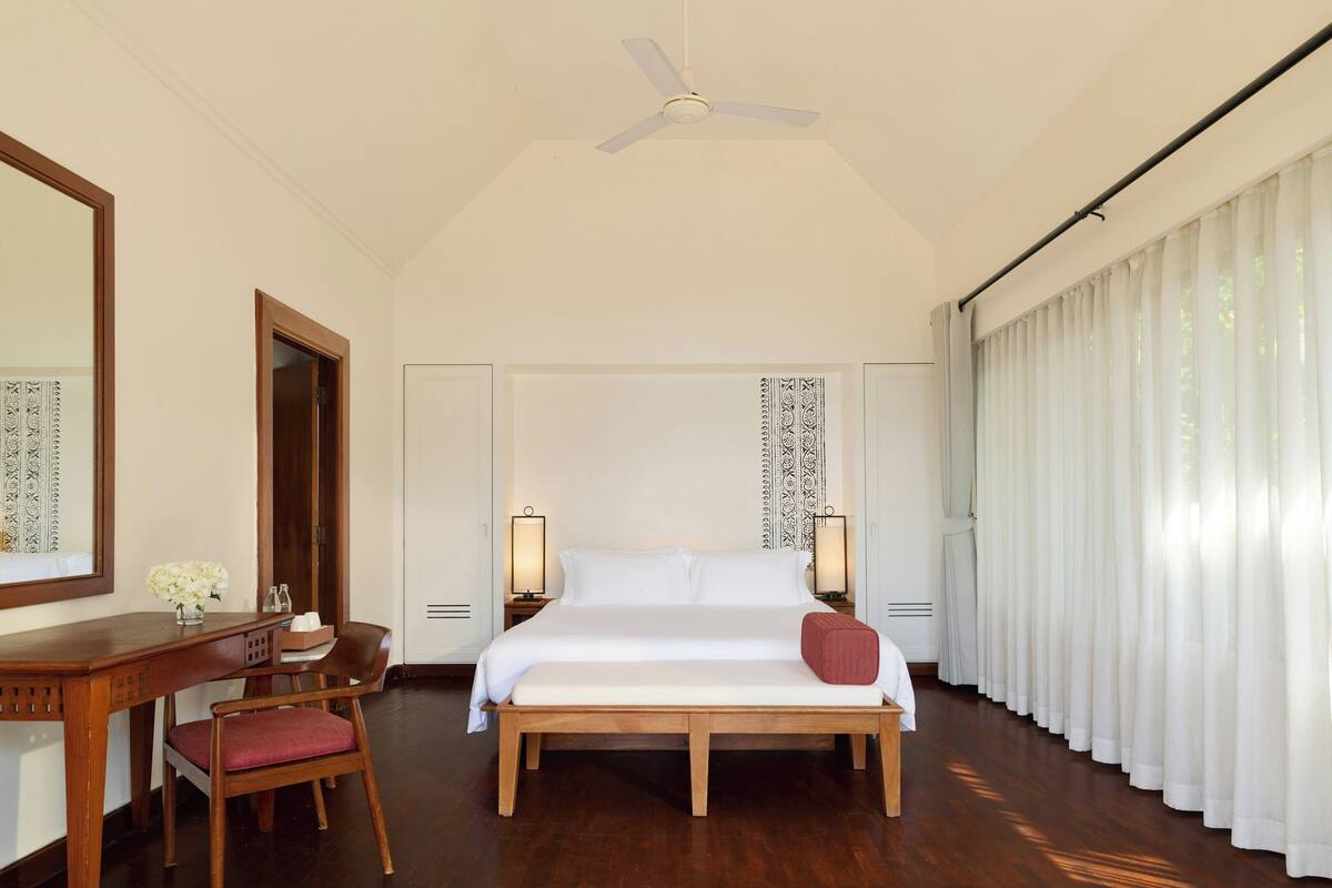 A hotel room with a bed, a desk, a chair, a ceiling fan, and a window with curtains.