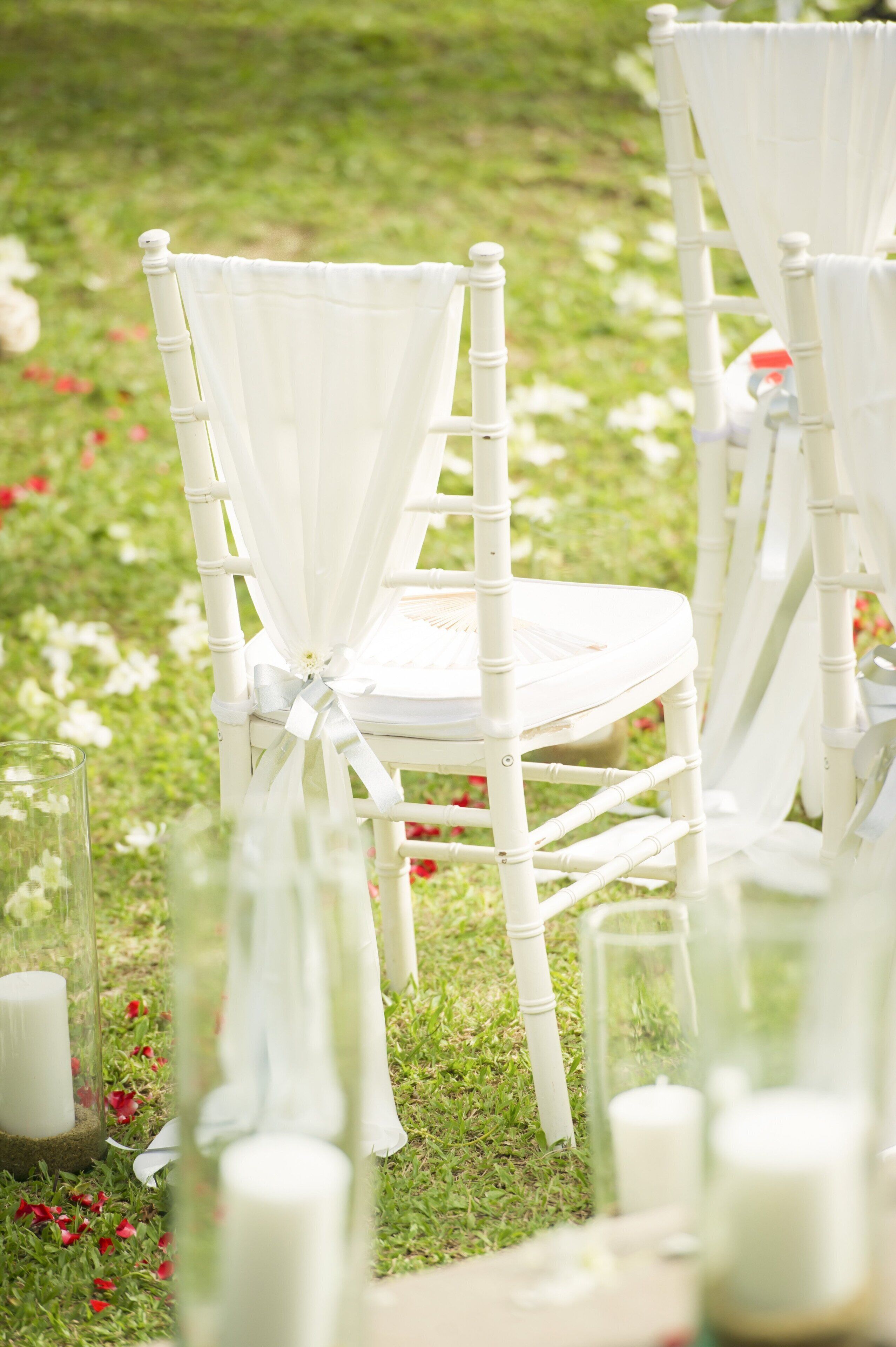 Une chaise blanche, ornée d’un ruban noué autour du dossier, est placée sur une pelouse parsemée de fleurs rouges.