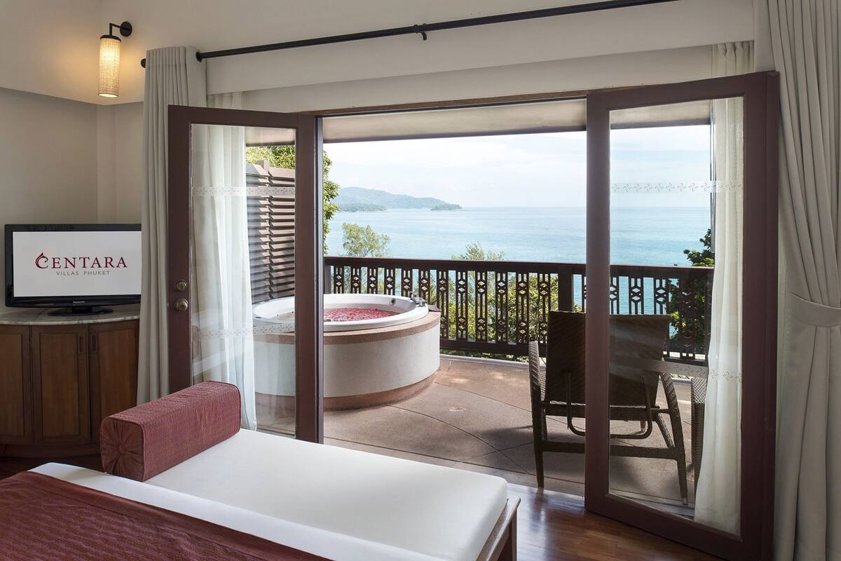 A hotel room with a bed, a TV, a hot tub, and a balcony with a view of the sea.