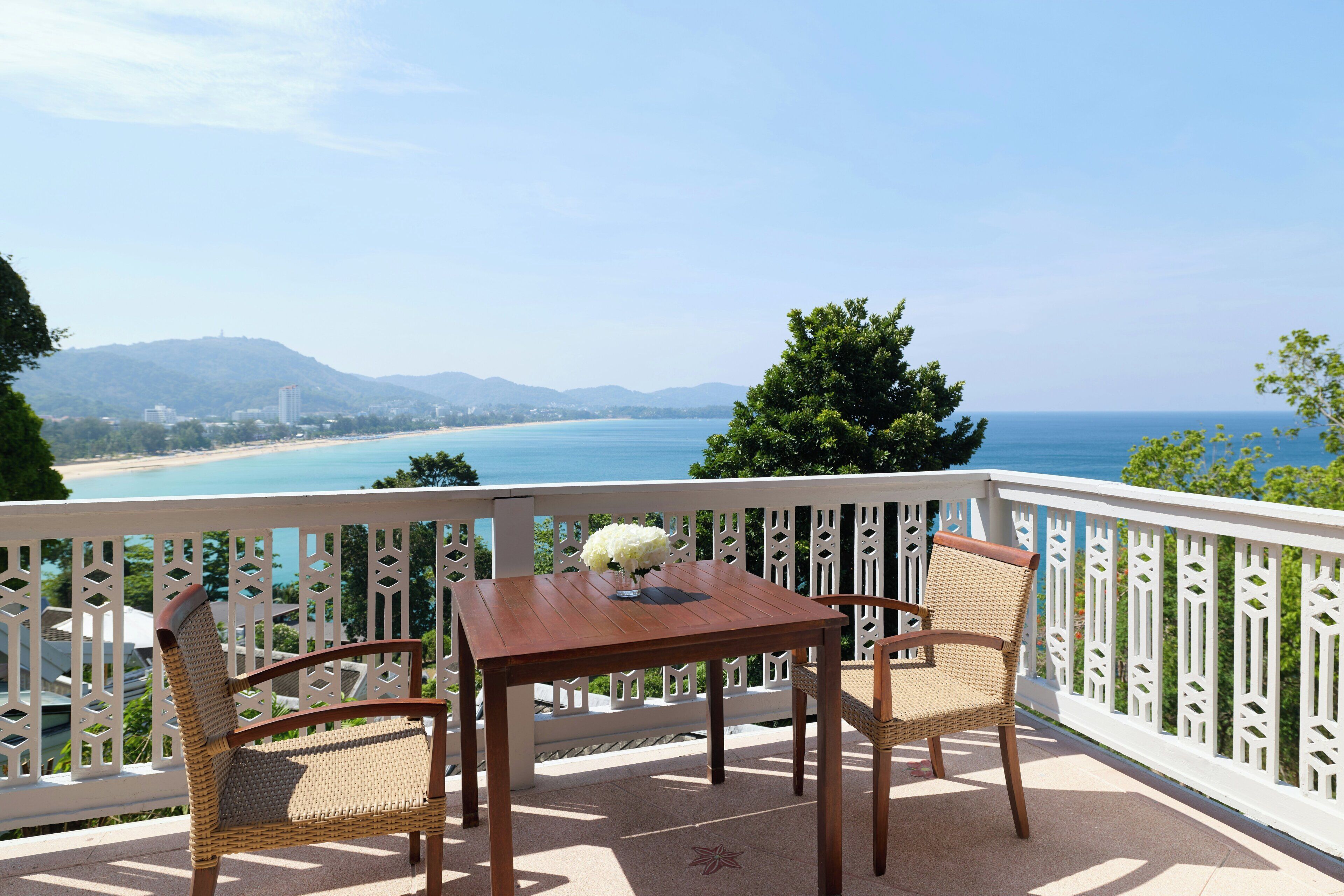 A balcony with a table and two wicker chairs, overlooking a beach and ocean.