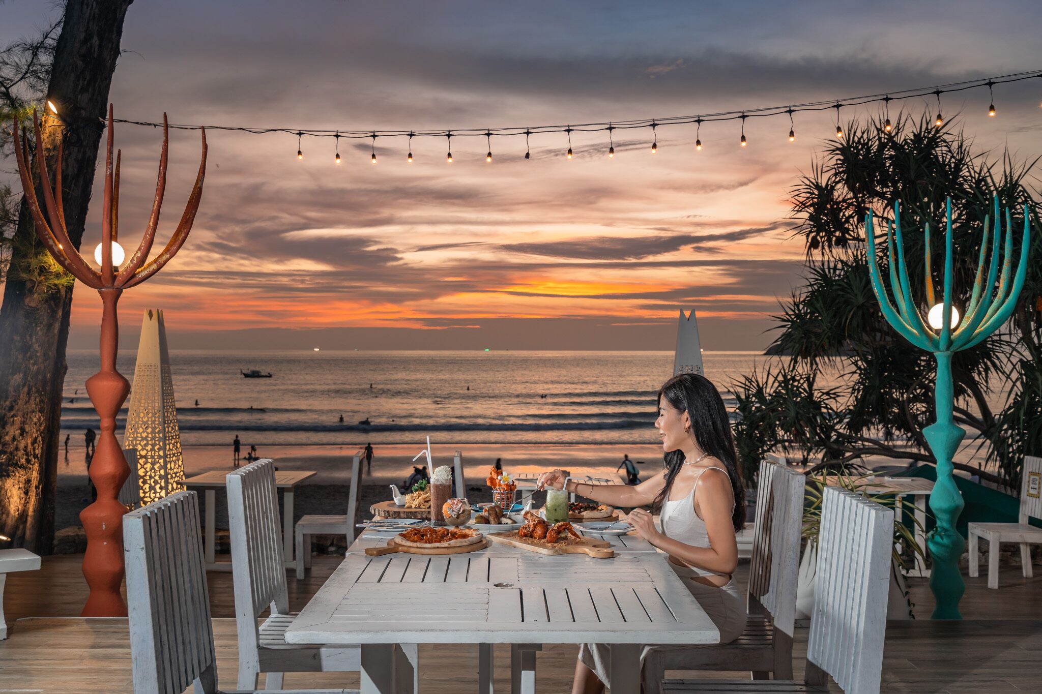 Une table à manger dressée avec de la nourriture et des boissons, donnant sur une plage au coucher du soleil.