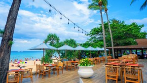 An outdoor dining area with wooden tables and chairs, umbrellas, and a view of the beach. - Beyond Kata (Karon)