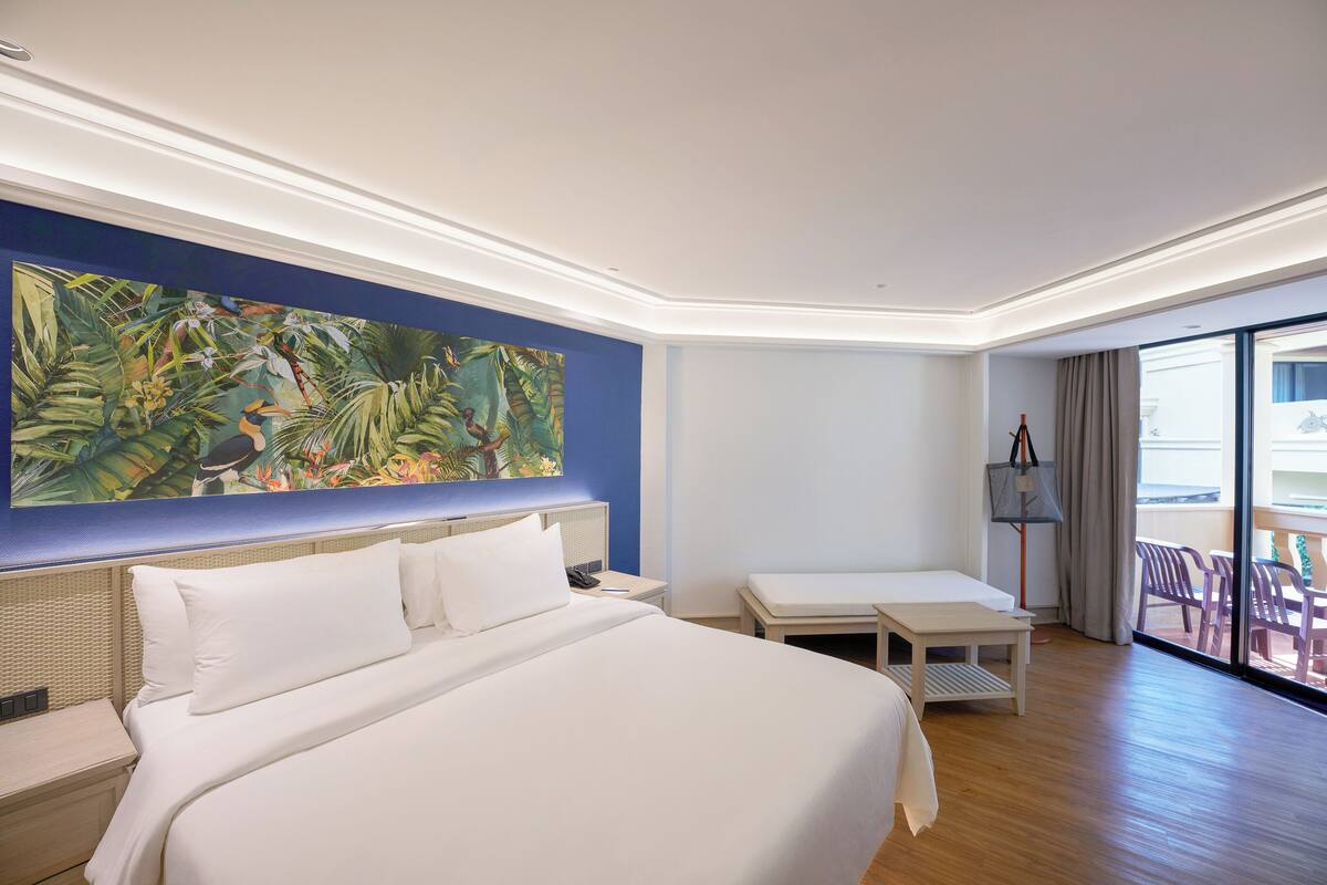 Une chambre d’hôtel avec un grand lit, une table de chevet, un bureau, une chaise, une grande fenêtre et un tableau aux motifs tropicaux accroché au mur.