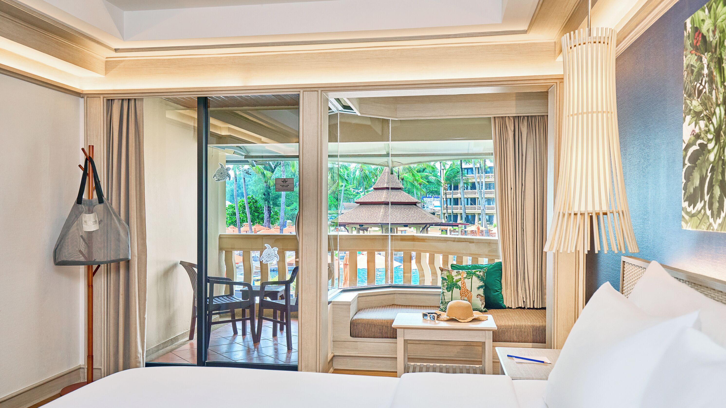 A hotel room with a bed, a desk, a chair, a balcony with a view of a pool and palm trees, and a hanging bag.