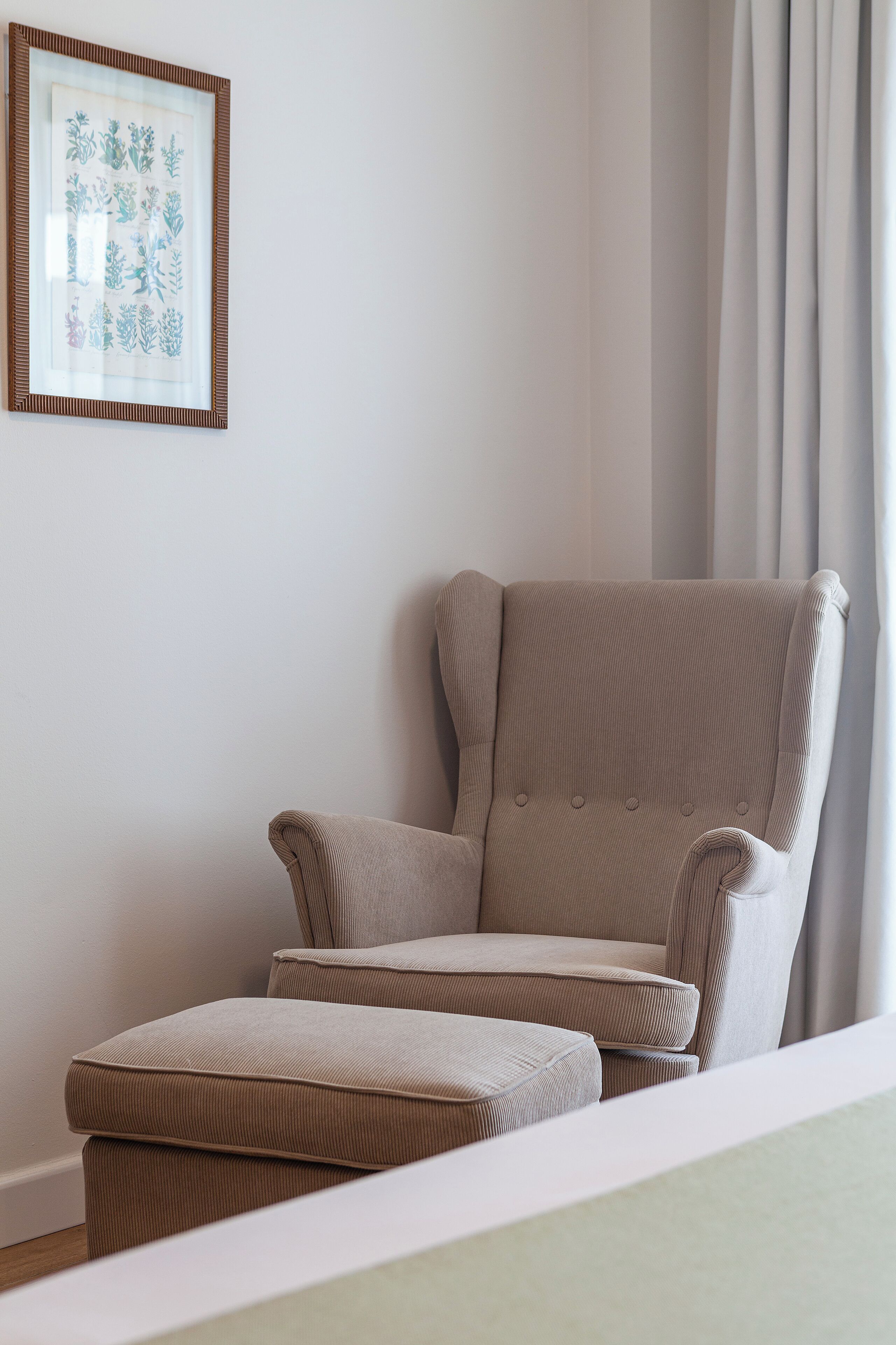 A corner of a room with a beige armchair and ottoman, a framed botanical print on the wall, and a window with curtains.