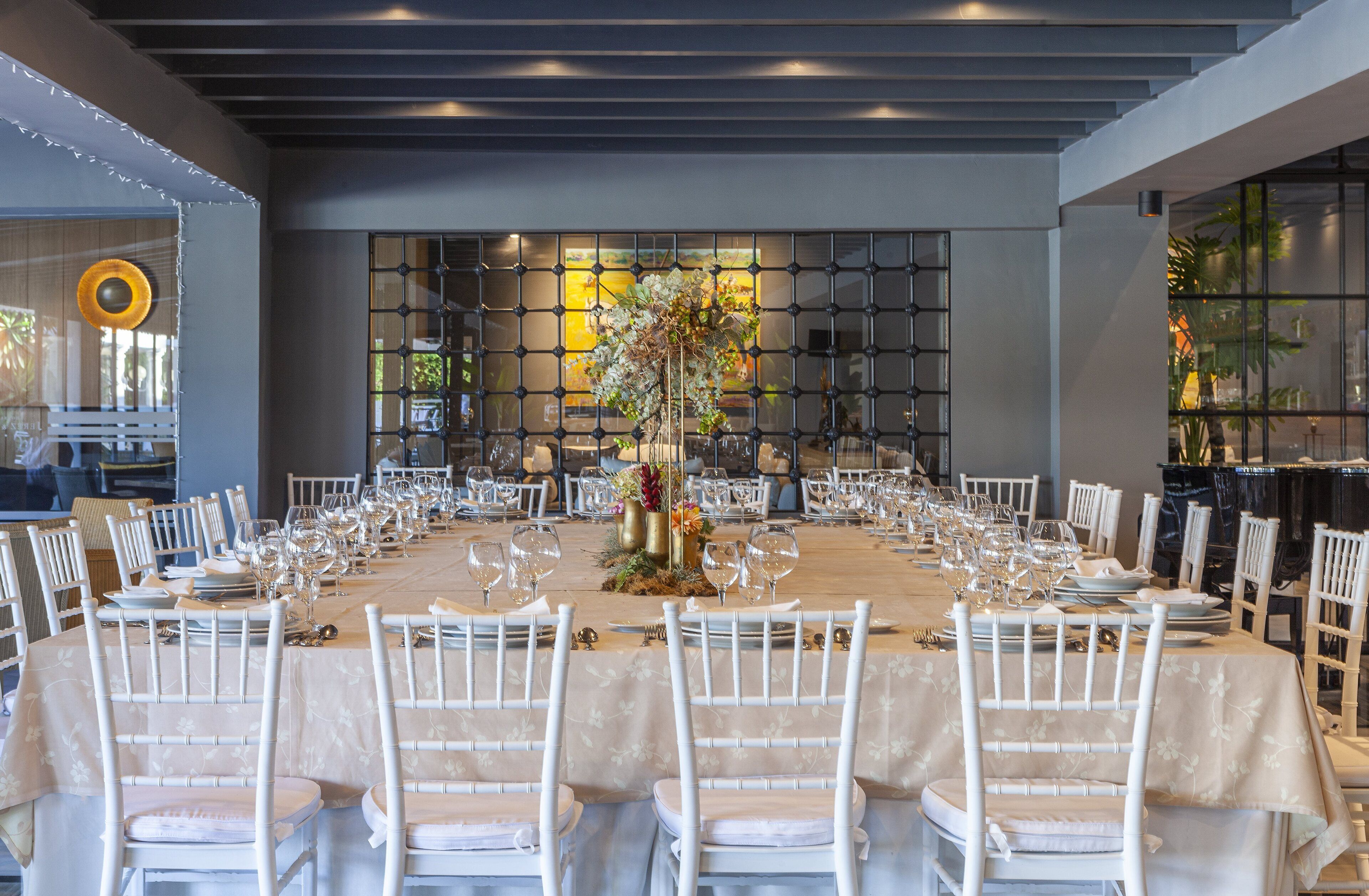 Una amplia mesa de comedor elegantemente dispuesta, con sillas blancas, un arreglo floral central y un diseño de interiores moderno.