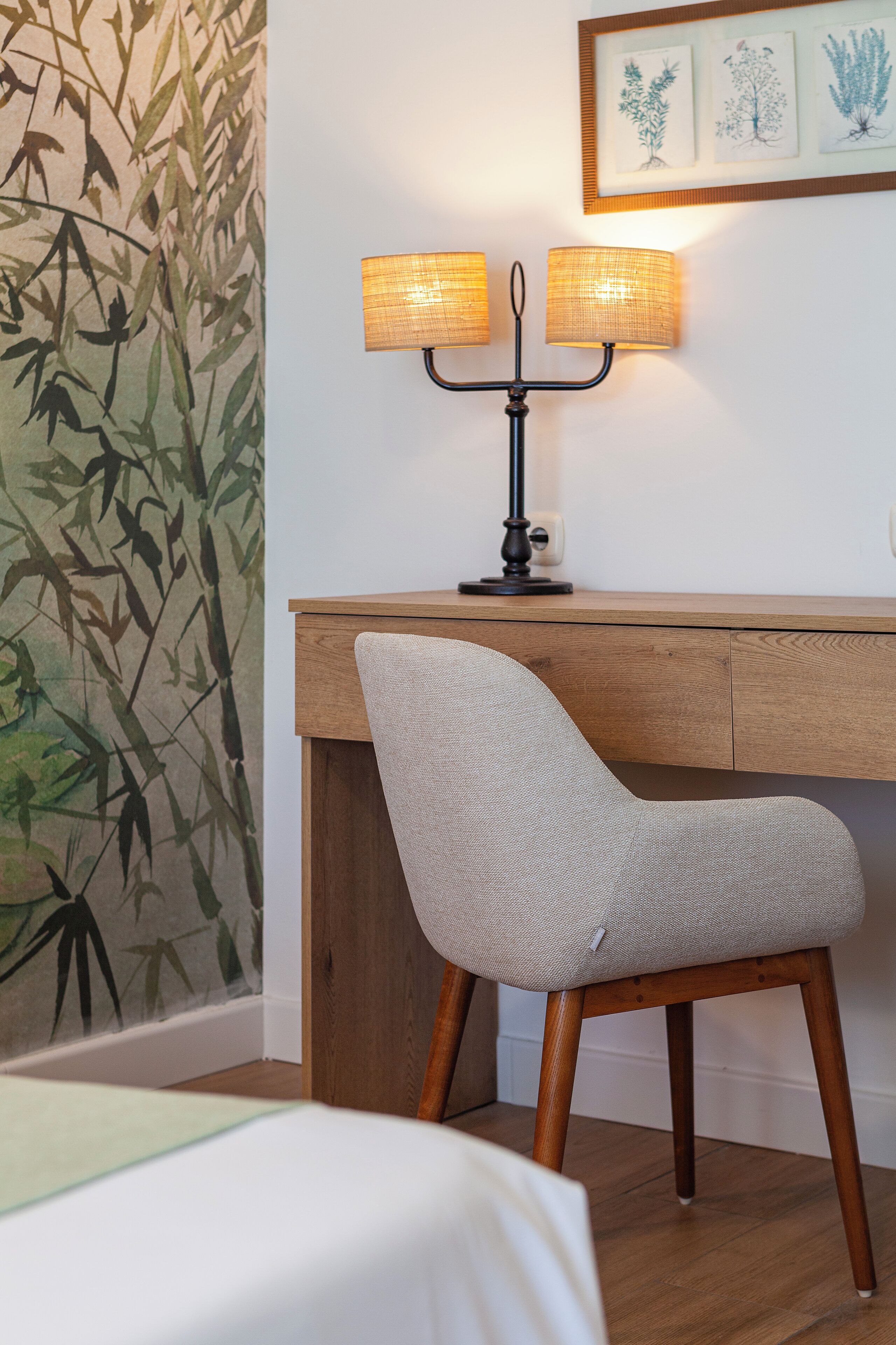 Une pièce avec un bureau en bois, deux lampes fixées au mur, une chaise et un mur recouvert d’un papier peint à motifs végétaux.