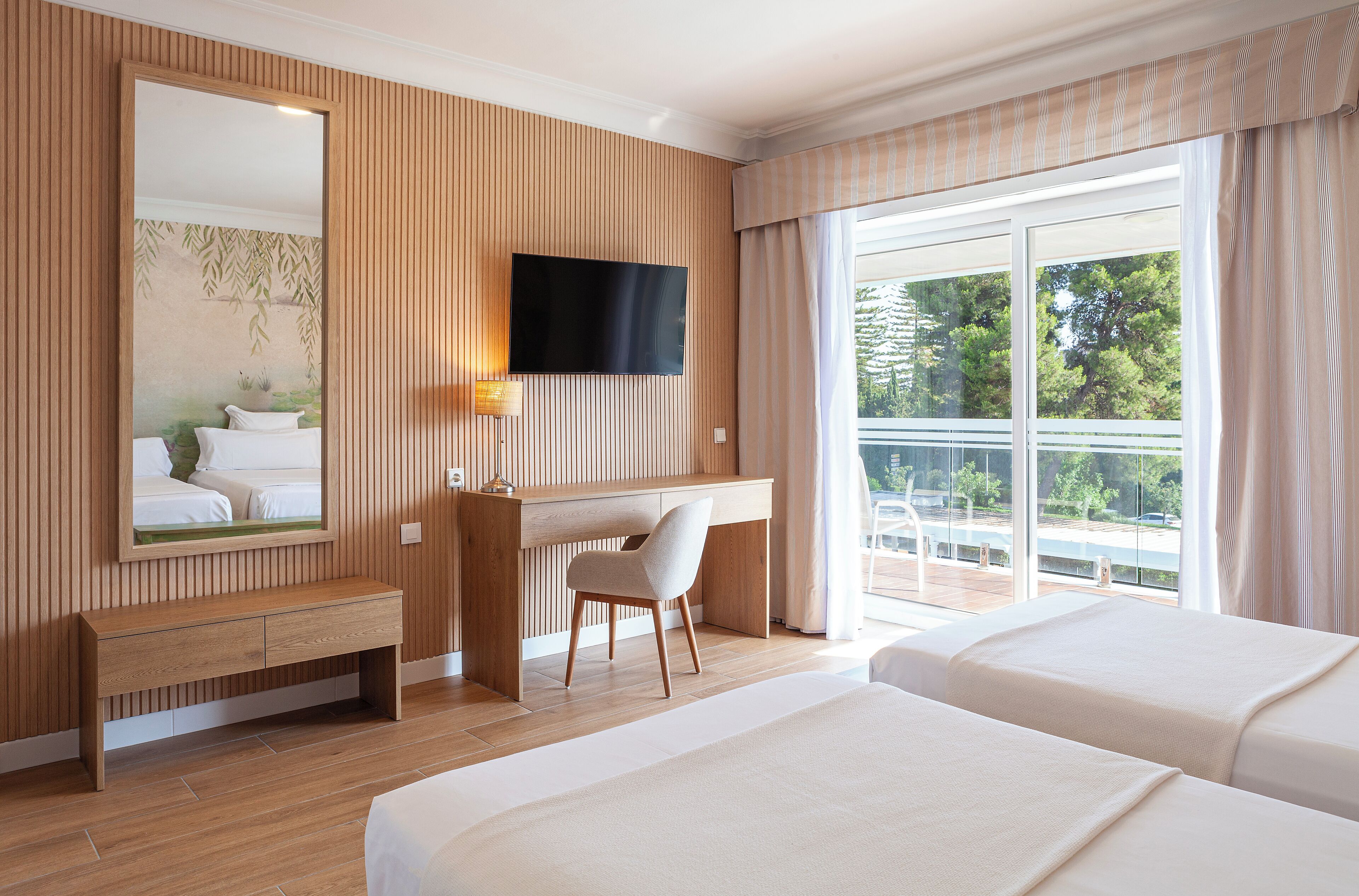 Quarto de hotel com parede revestida de madeira, um espelho grande, televisão de tela plana, escrivaninha com cadeira e duas camas.