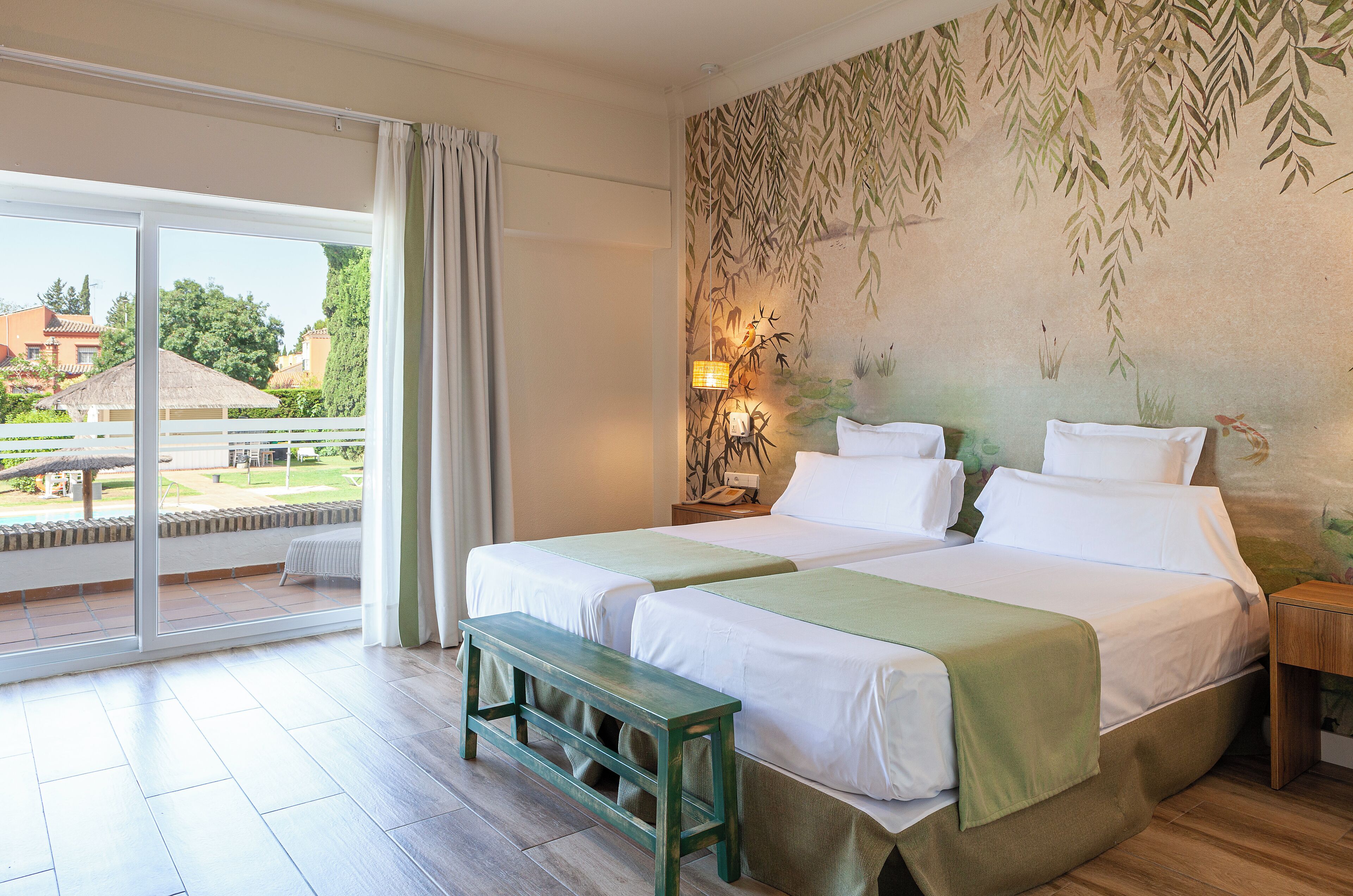 A double bed with a green bedspread, a bench, a nightstand with a lamp, and a view of a garden through a sliding glass door.