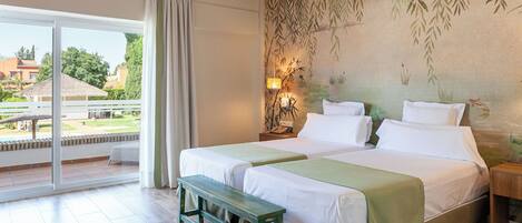 A double bed with a green bedspread, a bench, a nightstand with a lamp, and a view of a garden through a sliding glass door.