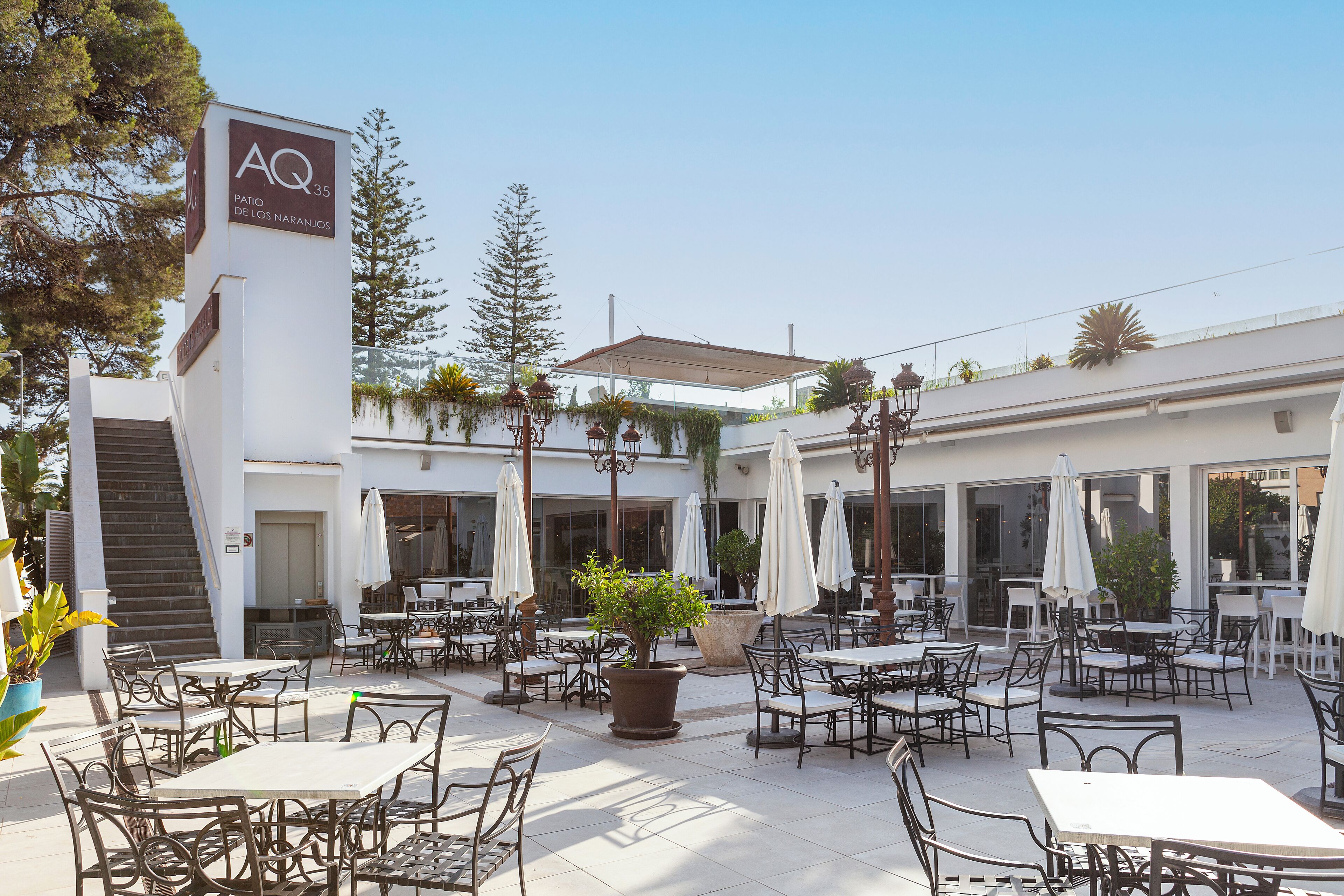 An outdoor cafe area with tables and chairs, a large planter with a tree, and a building with a sign reading 'AQ 35'.