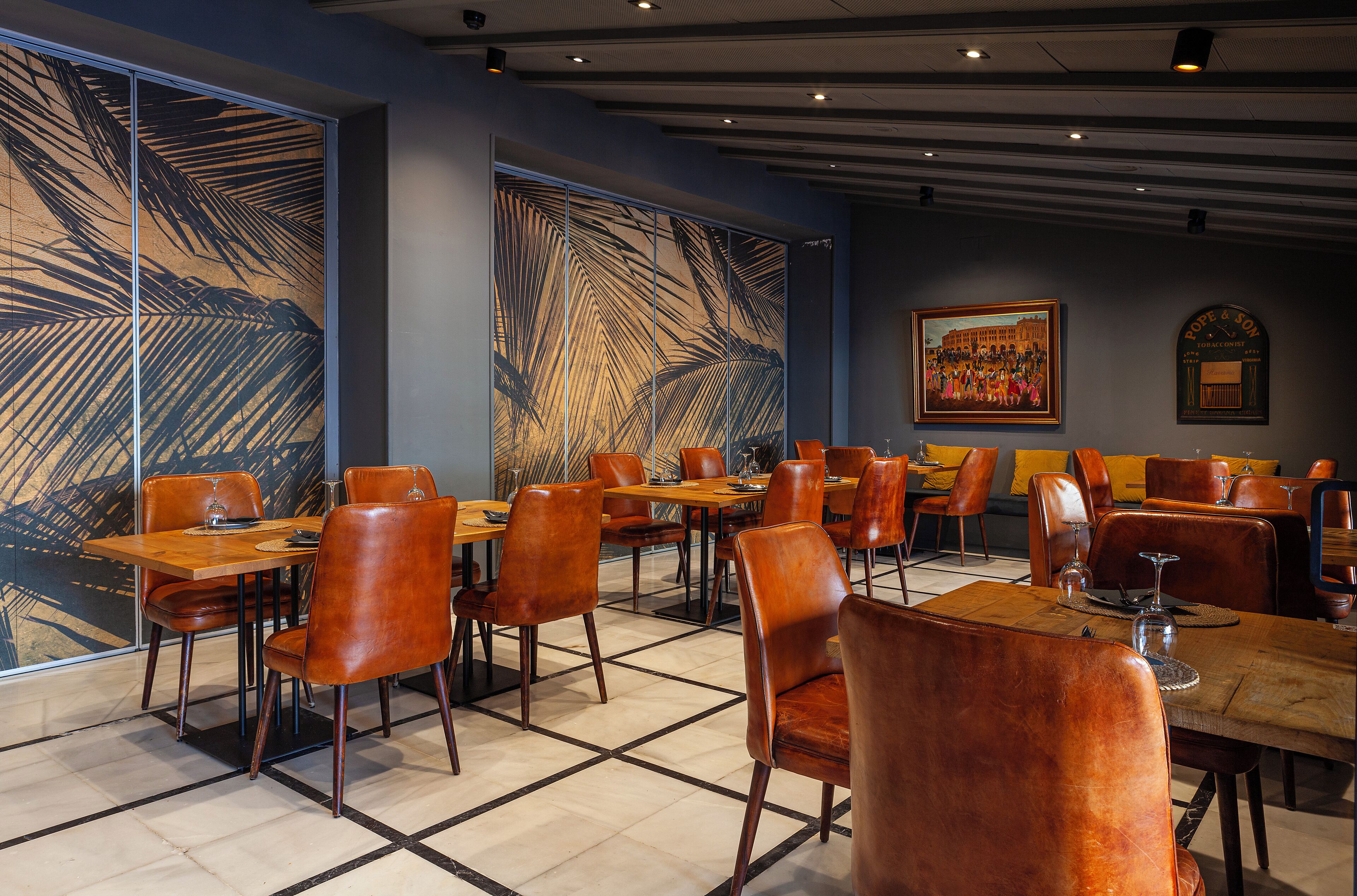 A restaurant with wooden tables and leather chairs, large windows with palm leaf patterns, and a framed painting on the wall.