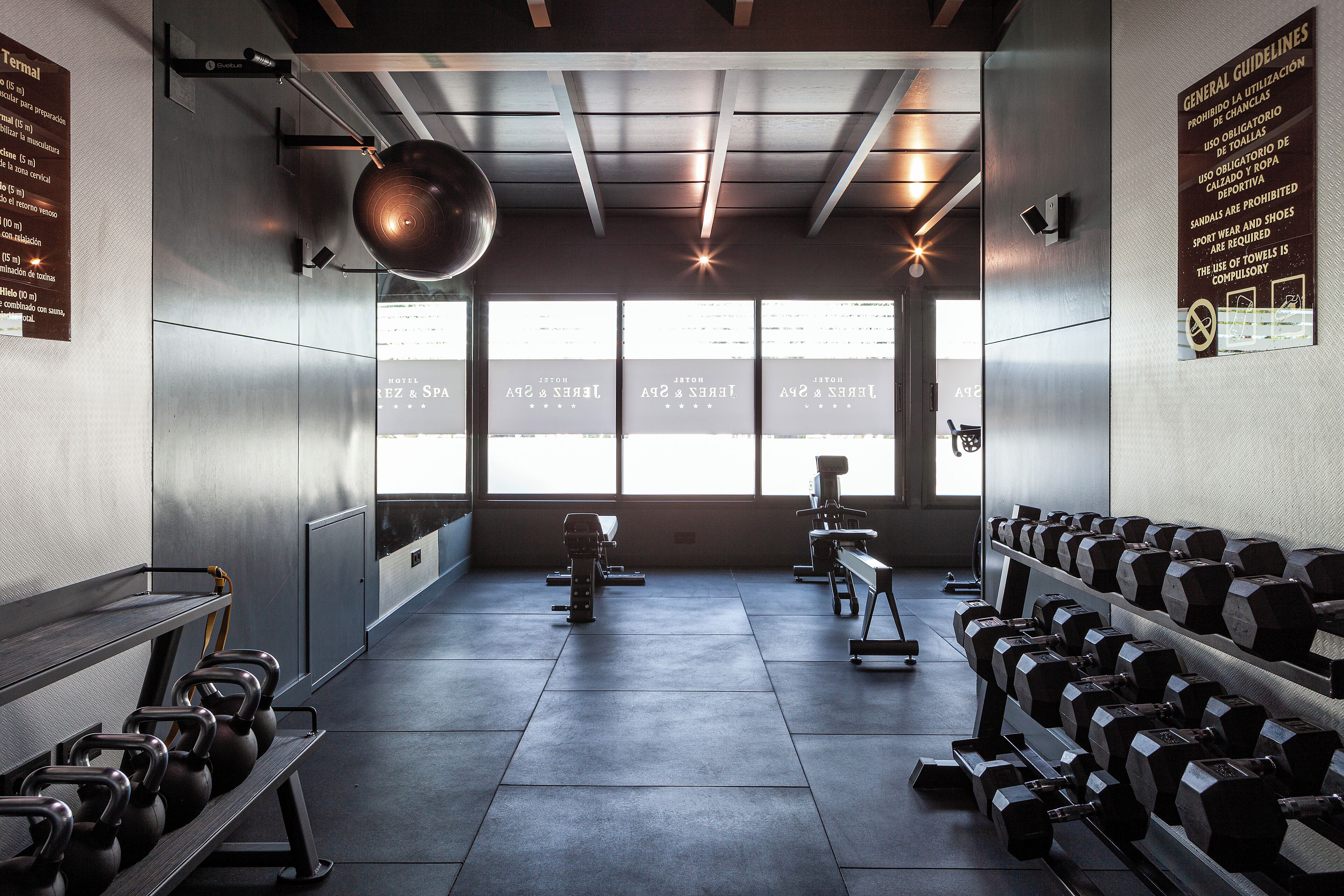 Un gimnasio con equipo de ejercicio, ventanas amplias y un cartel con pautas generales.
