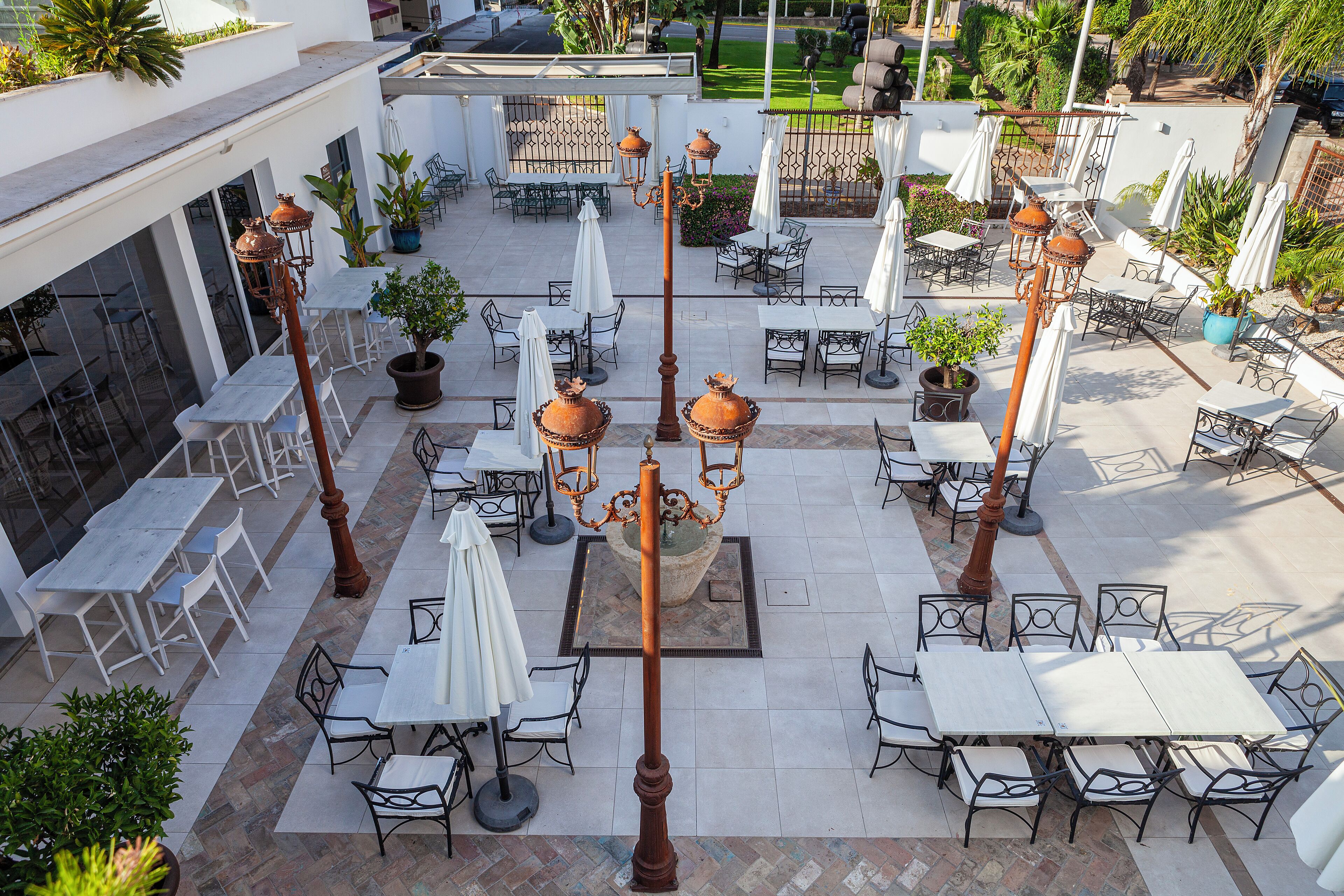 Een terras buiten met tafels en stoelen, omringd door potplanten en parasols.