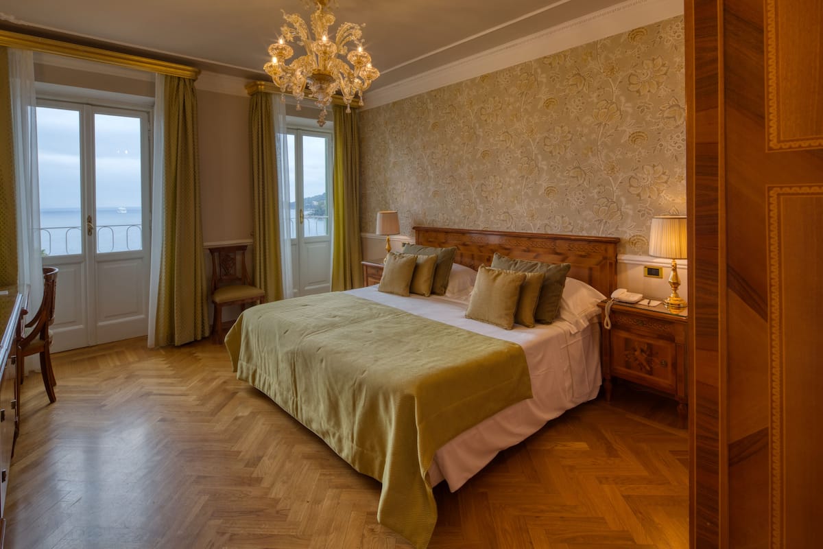 A spacious bedroom with a large bed, a wooden headboard, a chandelier, and a view of the sea through a balcony door.