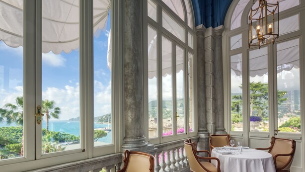 Un comedor con una mesa lista para dos, ventanas amplias con vista al mar y follaje, y sillas de madera clásicas.