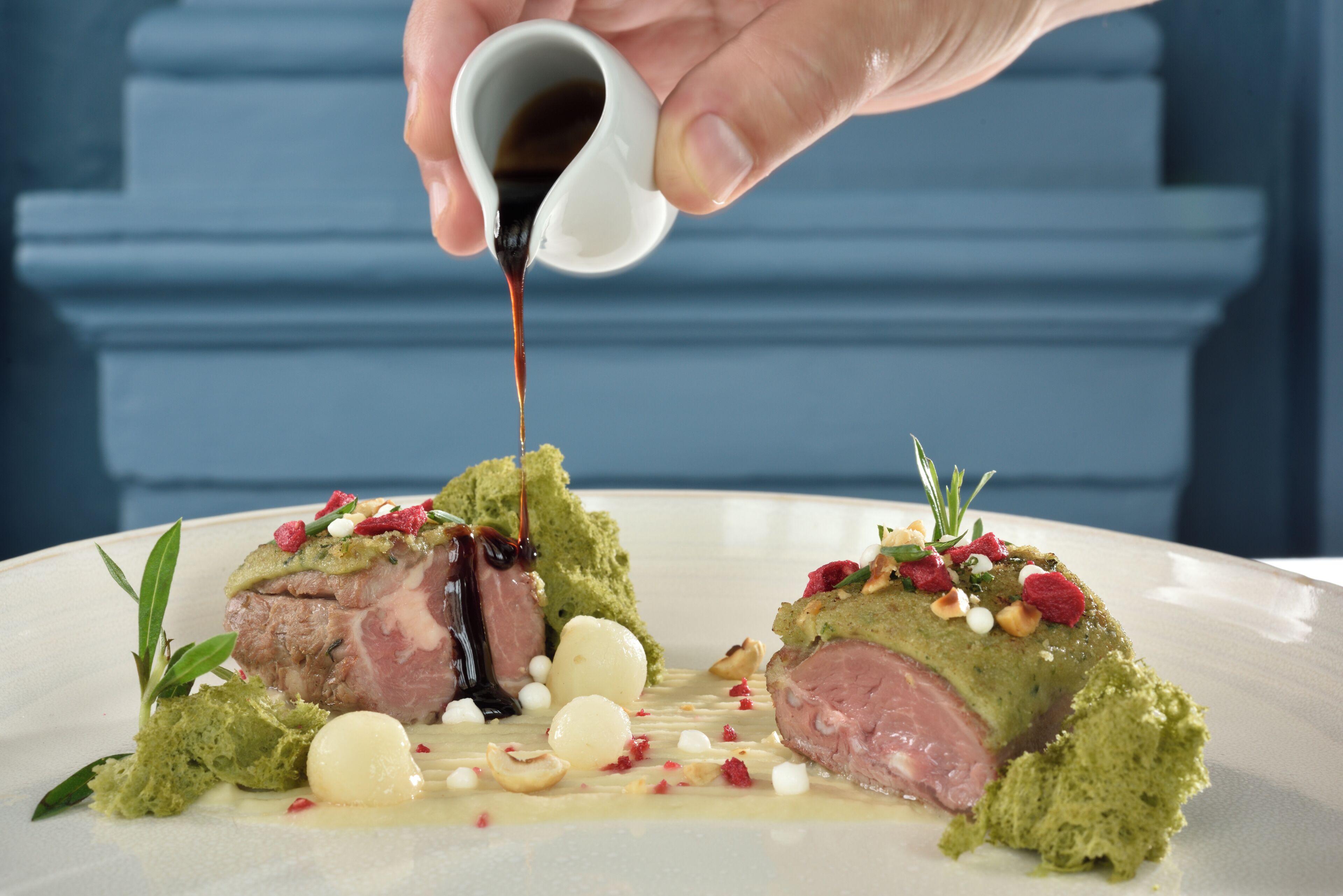 A hand pouring sauce over a dish with meat and garnishes.